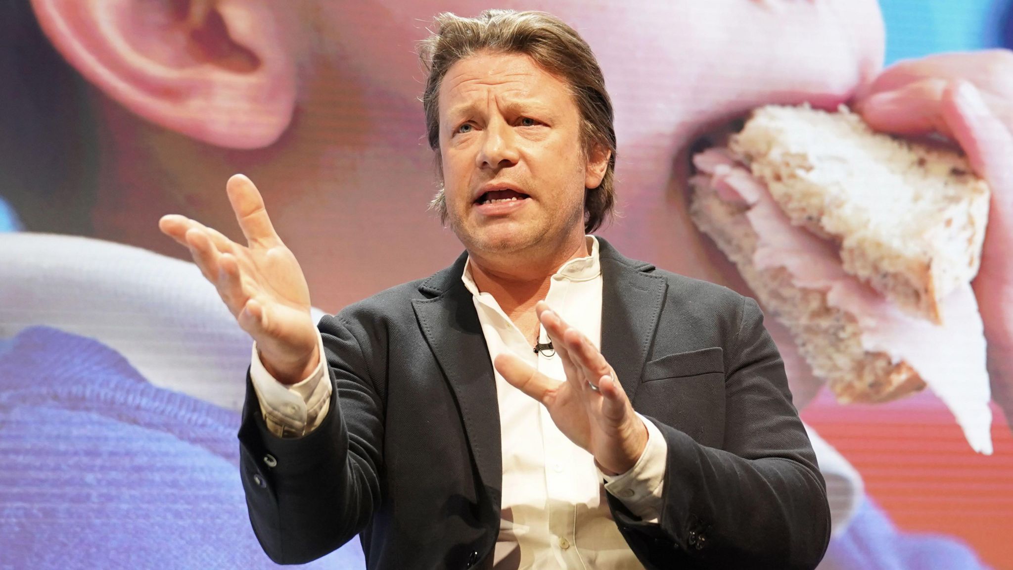 Jamie Oliver, sitting in front of a large poster of a child eating a sandwich,  speaking at the Tony Blair Institute for Global Change's Future of Britain Conference in London