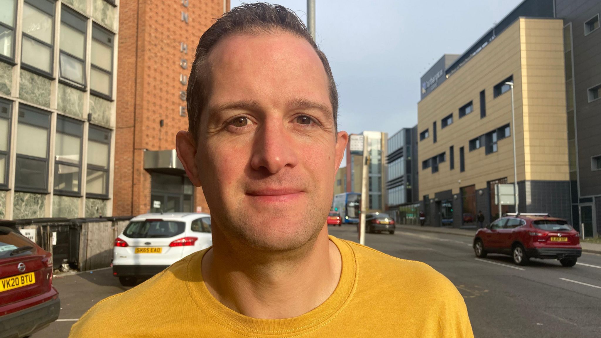 Rob Pardon smiles and looks directly at the camera wearing a yellow T-shirt. He is photographed outside on a pavement, with a street behind him. 