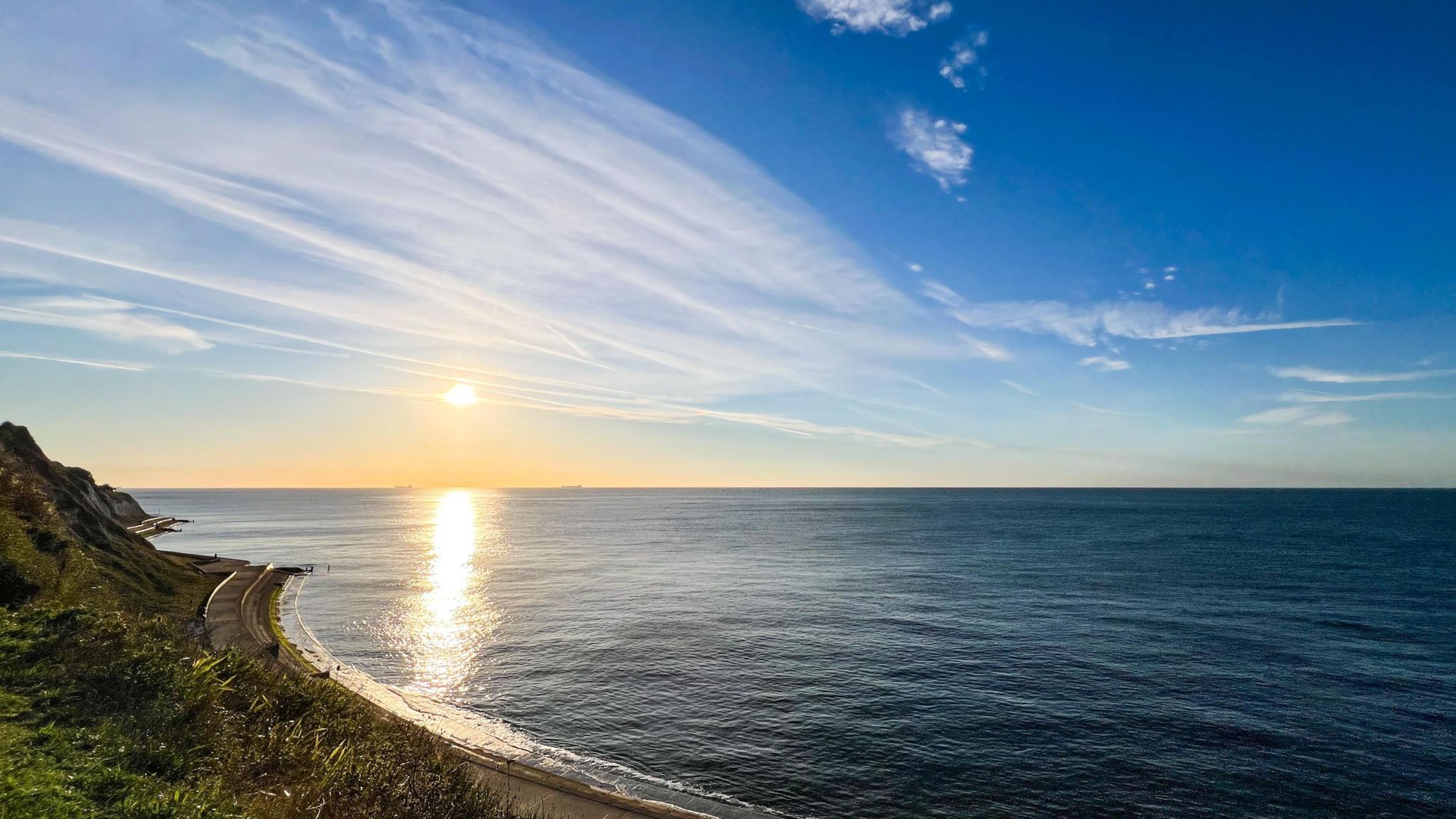 The dark blue sea stretches out to the horizon under a bright blue sky and with a beam of light from the sun stretching across the water 