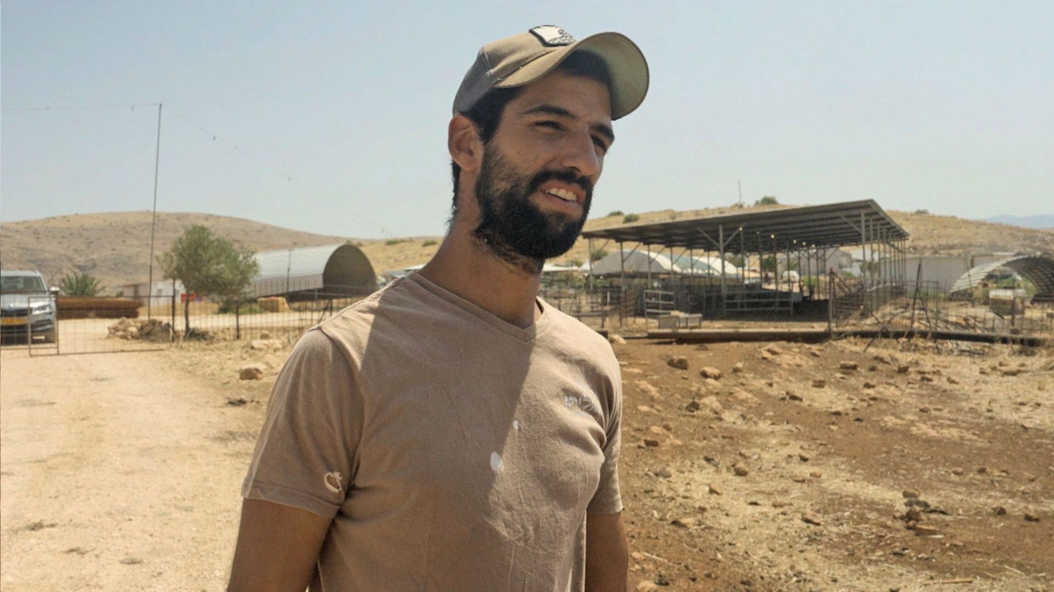 Moshe Sharvit, who has a beard and is wearing a khaki t-shirt and cap, captured in conversation with our interviewer. Behind him are metal constructions on his outpost and dusty-looking land, with a car parked behind a metal fence.