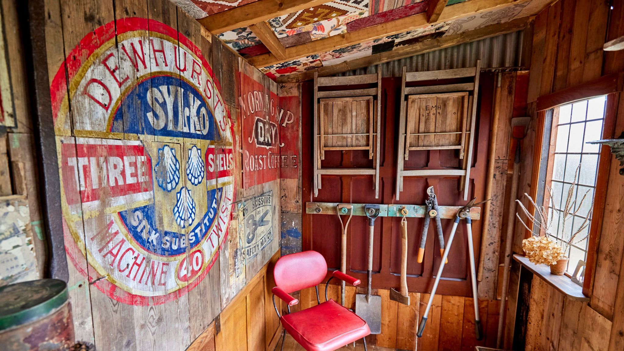 The interior of a shed with a red chair in the corner and gardening tools hung on the wall and there is a small window