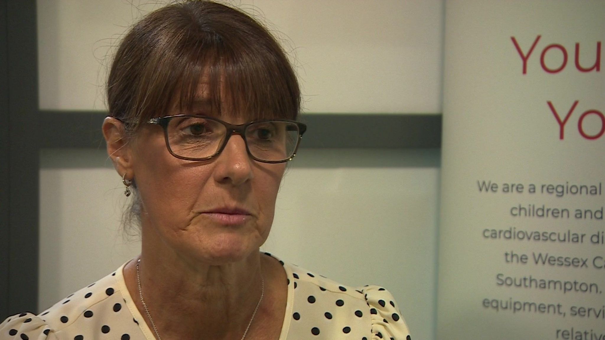 Sharon Brady wearing spotted dress and glasses, sitting in front of white background.