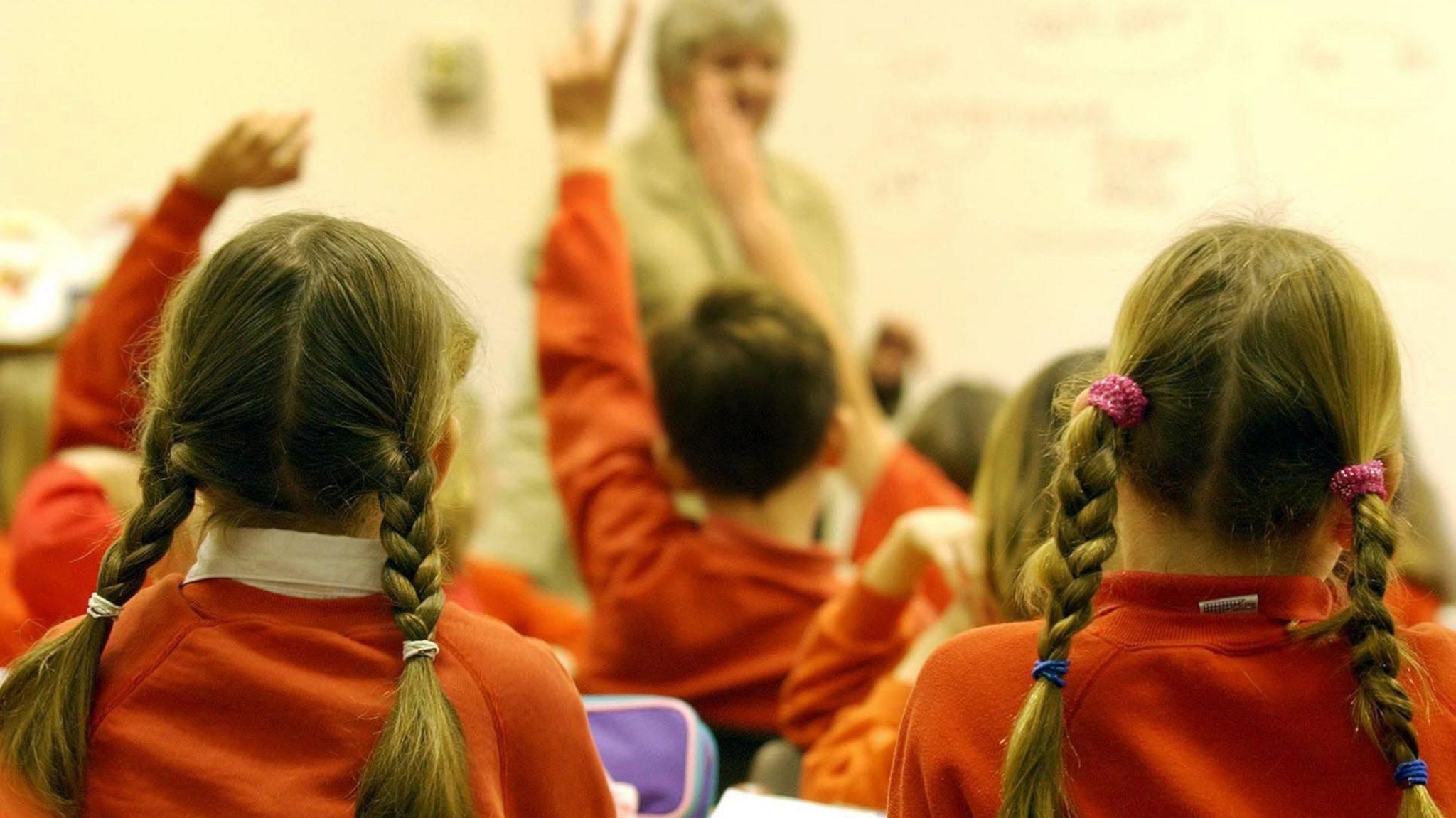 A view from behind of children in a classroom wearing red jumpers. Some have their hands raised to get the attention of their teacher who is in the foreground.