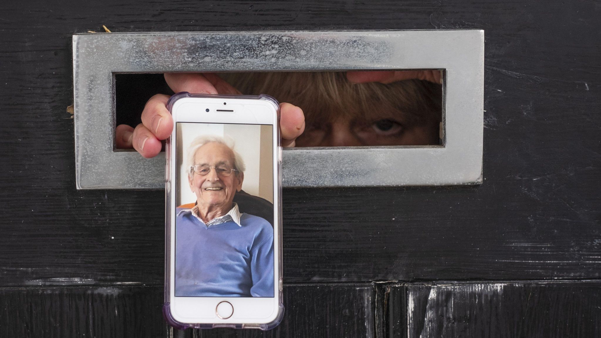 A person with a long blonde fringe looks through a letter box on a black door. They are pushing a smartphone through the letter box. On the phone there is what could be either a picture or video call of an elderly man wearing a cornflour blue jumper and a white shirt. He is smiling. 