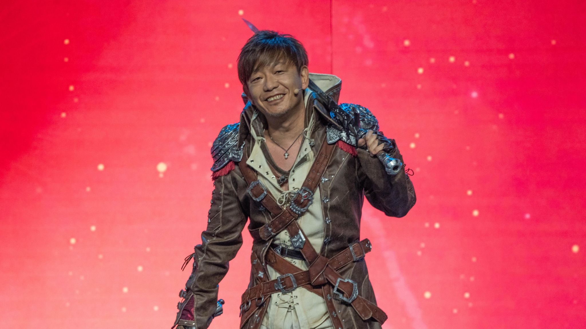 A man dressed in costume poses on a stage, smiling. He has a replica sword resting on the shoulder of his long leather jacket, which is fastened by several interlocking belts. The belts have ornate buckles that complement the intricate metal shoulder pads of the garment.