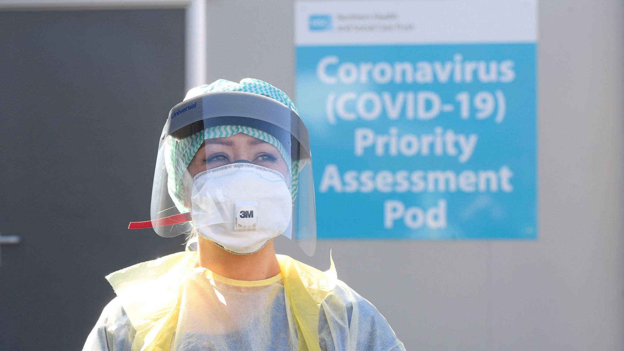 Woman wearing face covering and shield, standing in front of prefab building with sign reading 'Coronavirus (COVID-19) Priority Assessment Pod' 