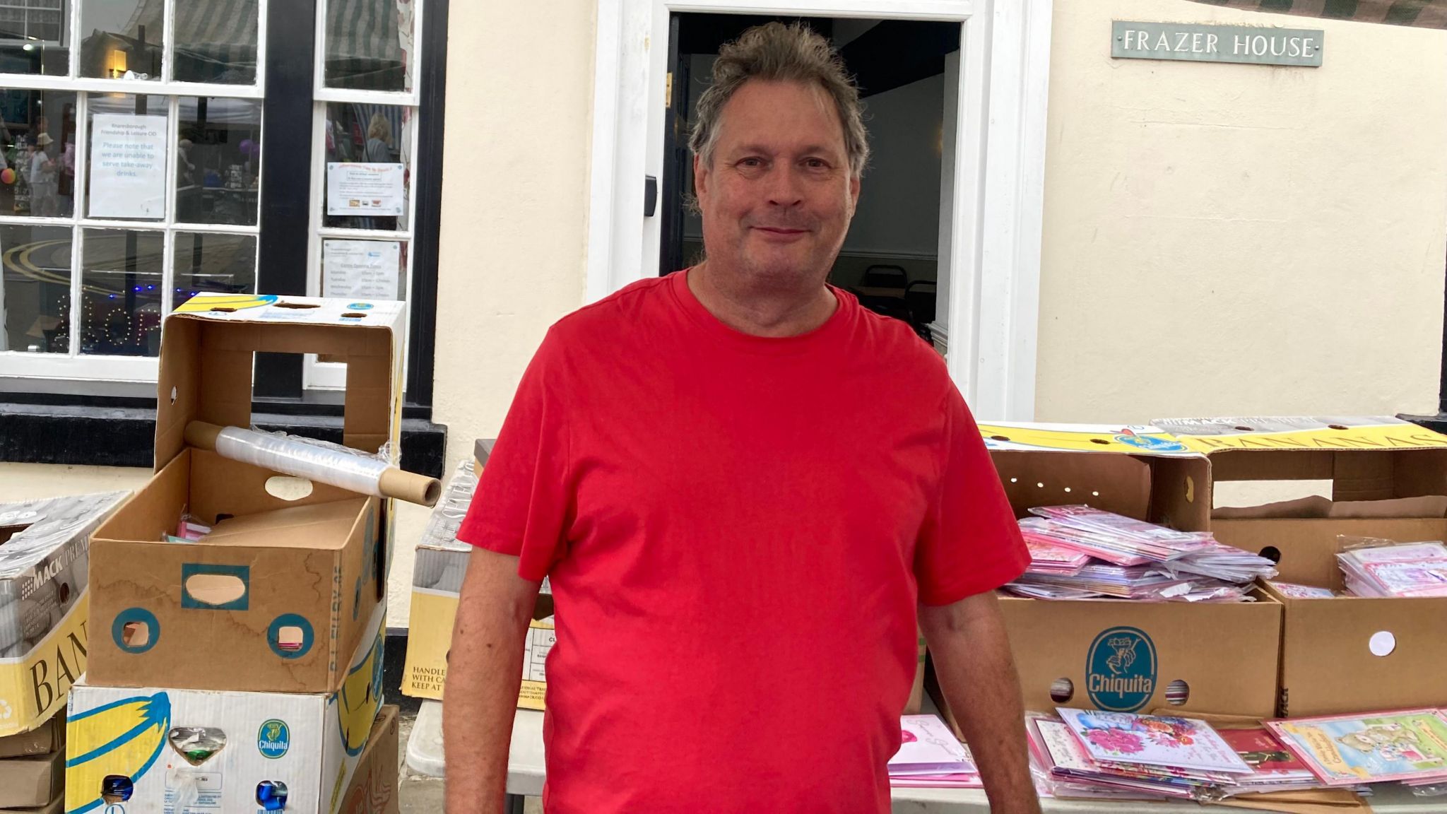 Image shows man wearing a red T-shirt surrounded by boxes of greeting cards