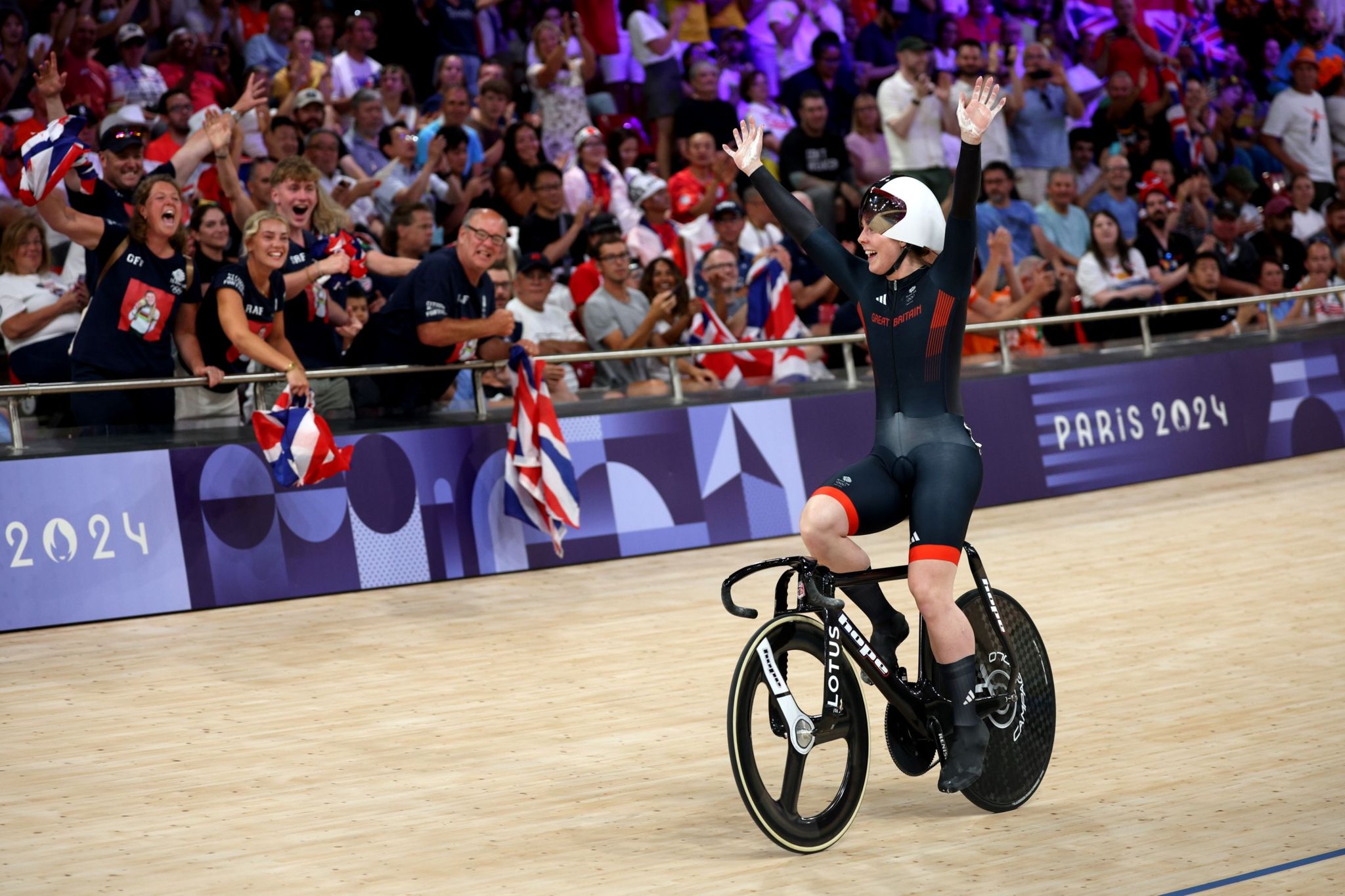 Emma Finucane sits on her bike with her hands raised, waving to the crowd of Team GB fans behind her. She is wearing a dark blue Great Britain cycling suit and has  her white helmet pushed up onto her head.