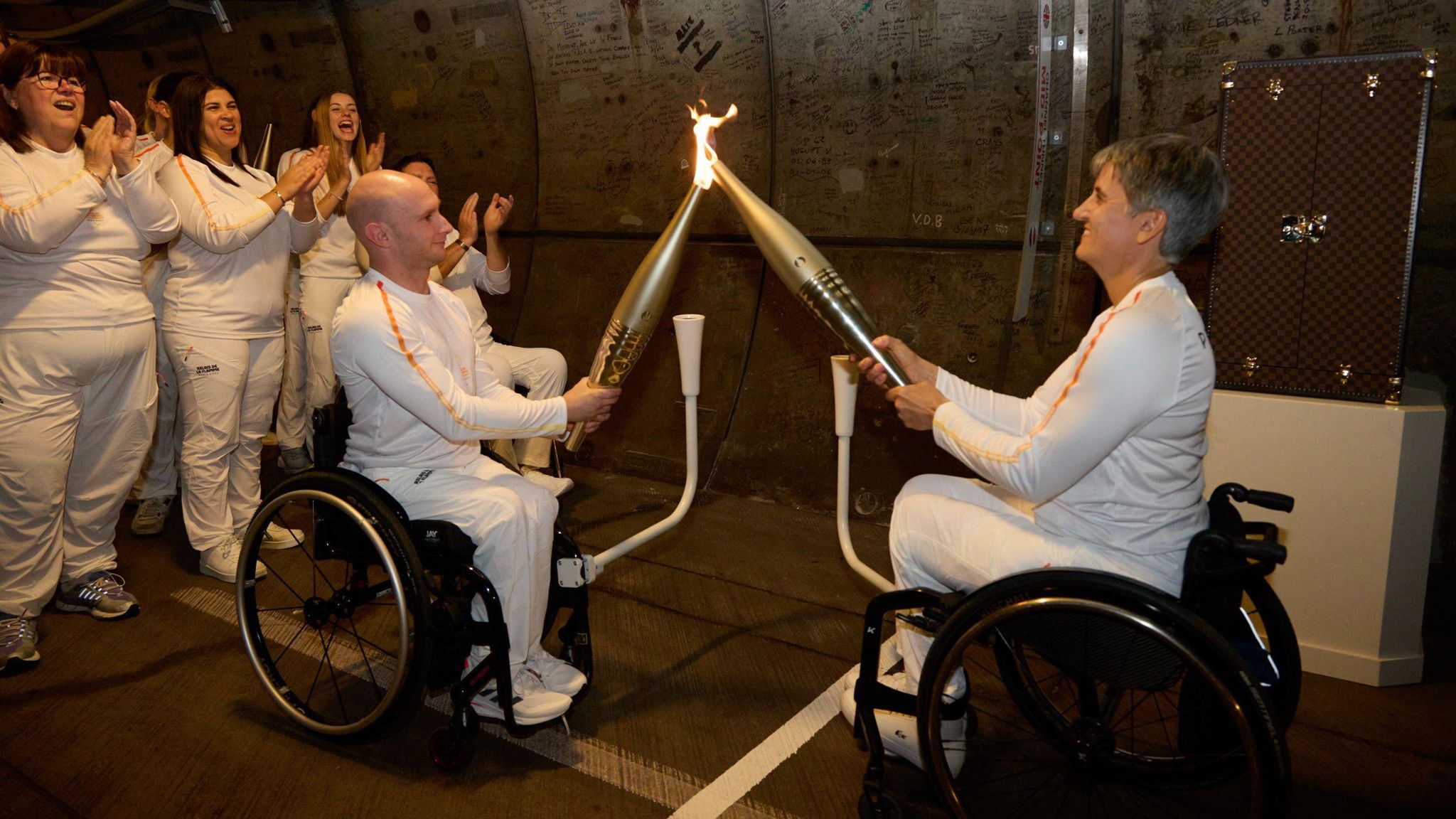 The Paralympic flame is handed over by two athletes who are wheelchair users while behind them a group is applauding