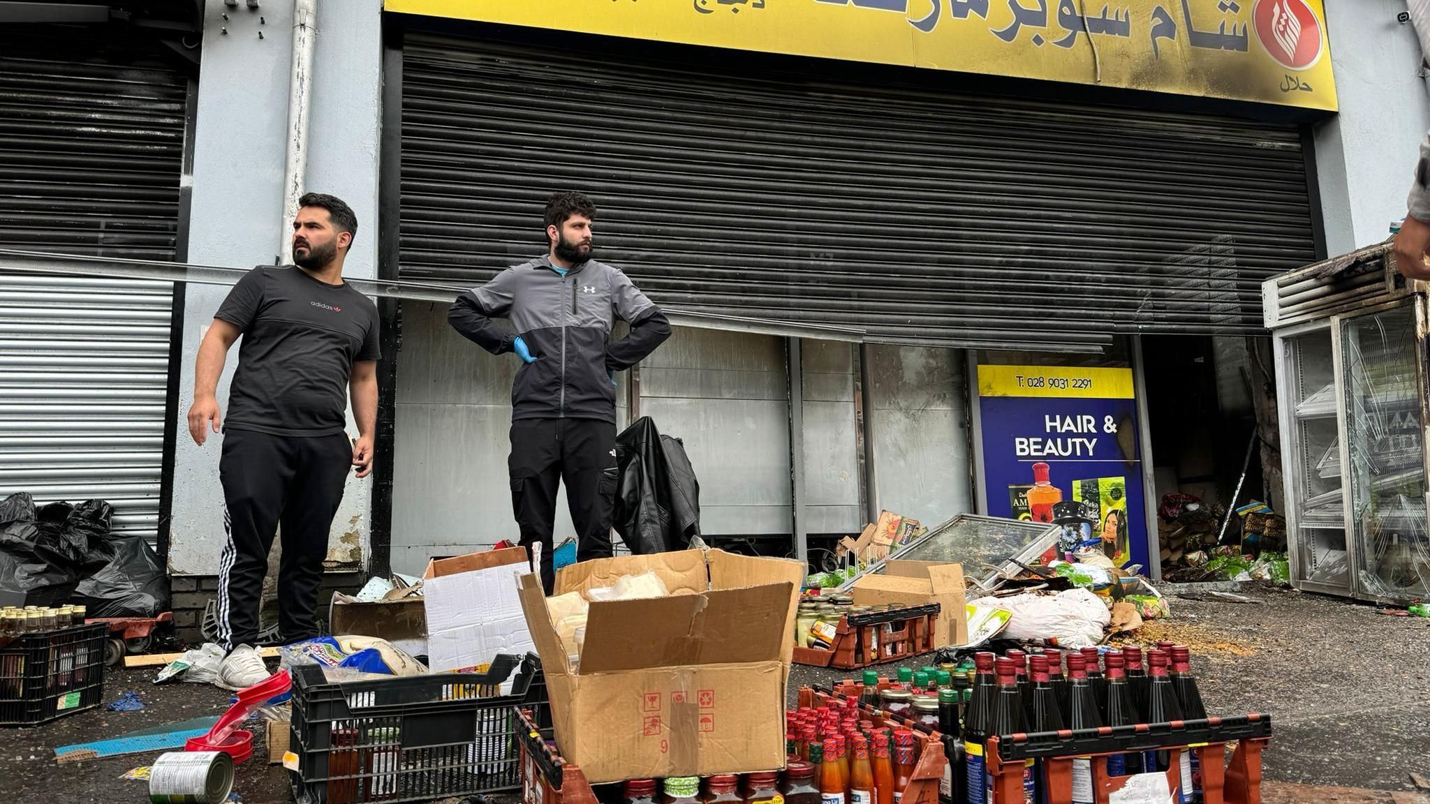 Two shopkeepers outside fire-damaged store on Donegall Road with damaged stock strewn across the pavement