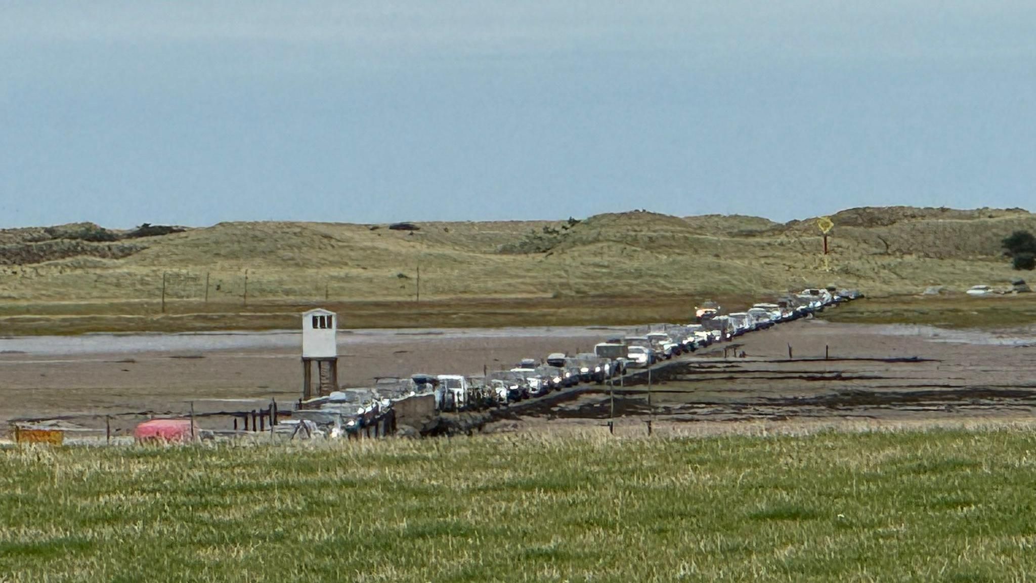 A queue of cars on a road surrounded by sand. Sand dunes can be seen in the background.