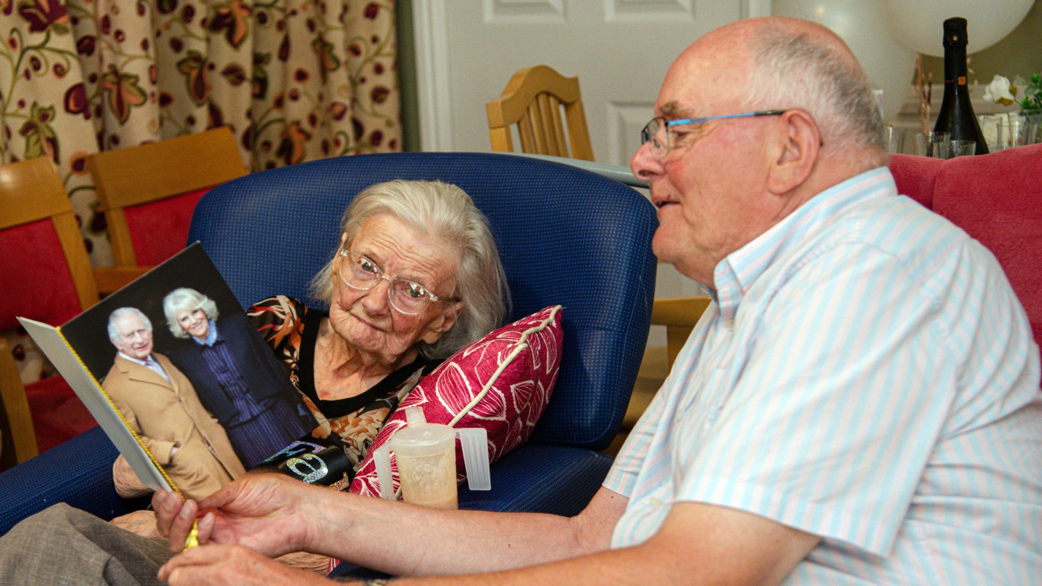 An elderly woman sat in a blue chair next to a man in a white and blue shirt, as they look at a card with King Charles and Queen Camilla on the front