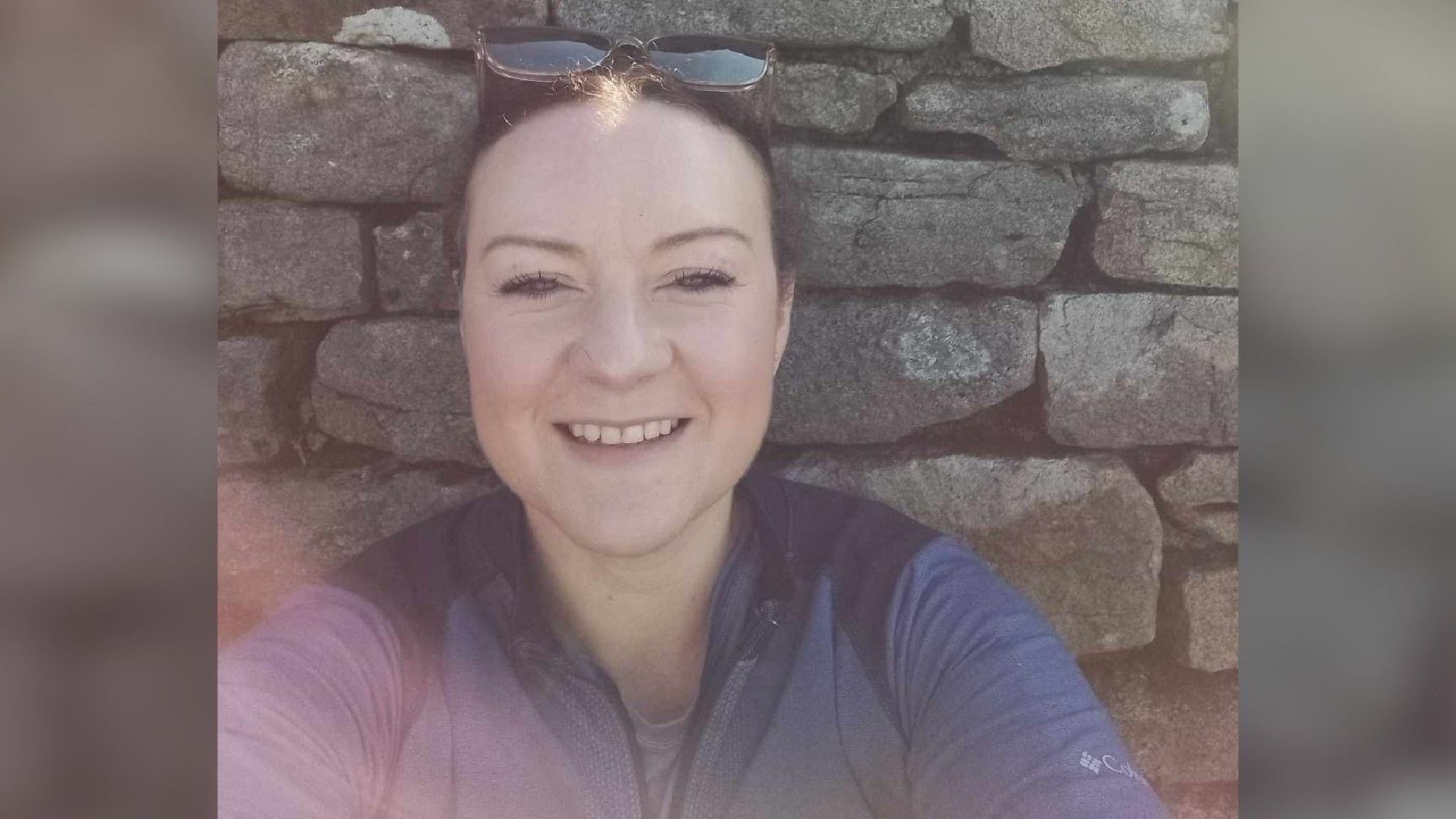 A woman in a dark coloured top with sunglass on top of her head smiles at the camera as she sits in front of a dry stone wall