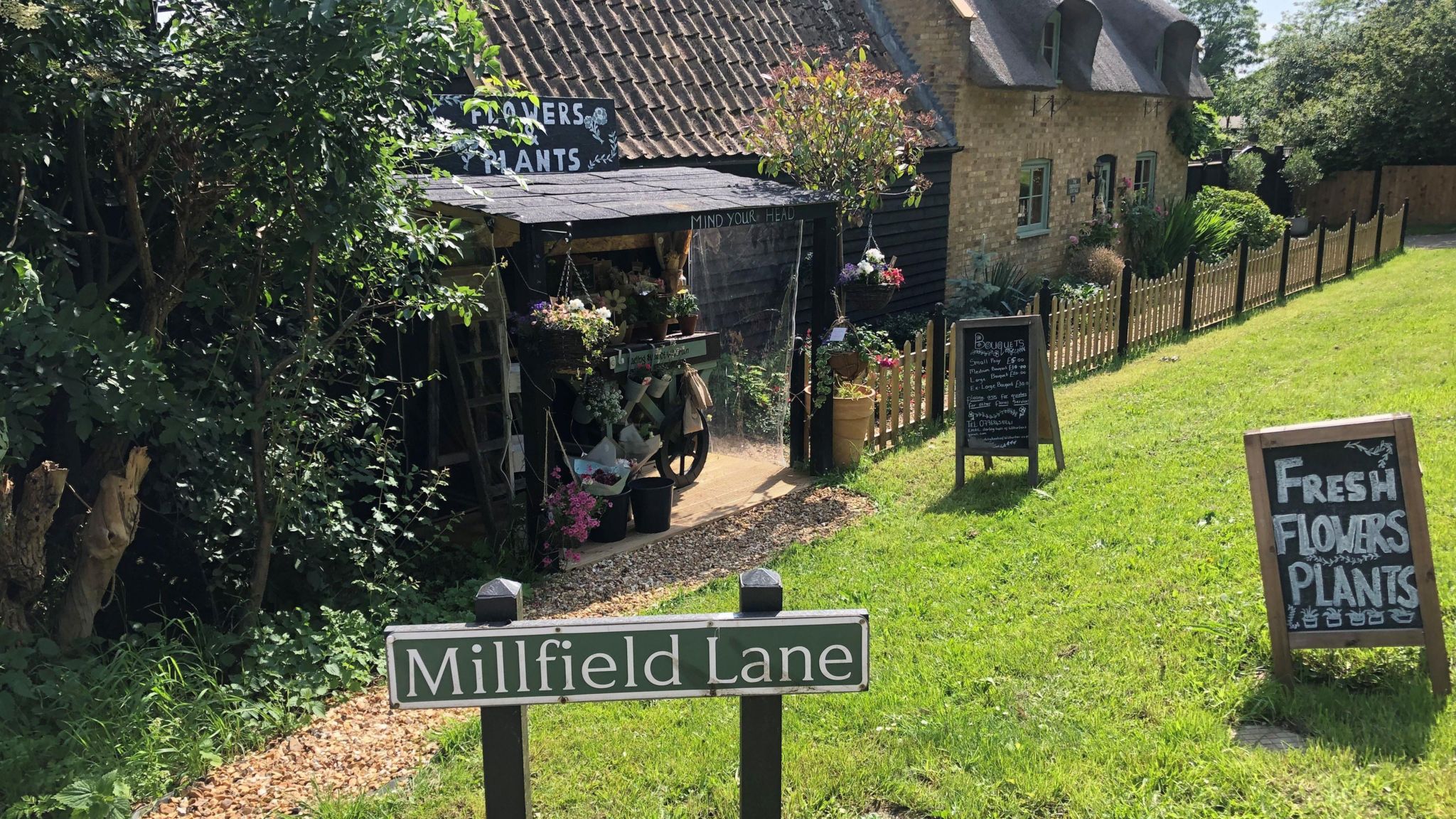 A flower stall outside a cottage behind a sign reading Millfield Lane