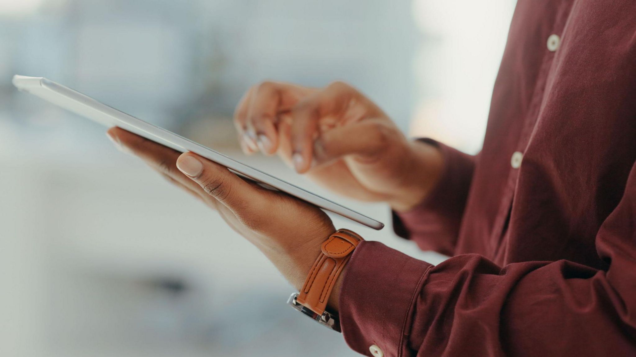 A person in a maroon red shirt is holding a tablet and is tapping on the screen. They are wearing a brown watch. You can only see part of their torso, and the background is blurred.