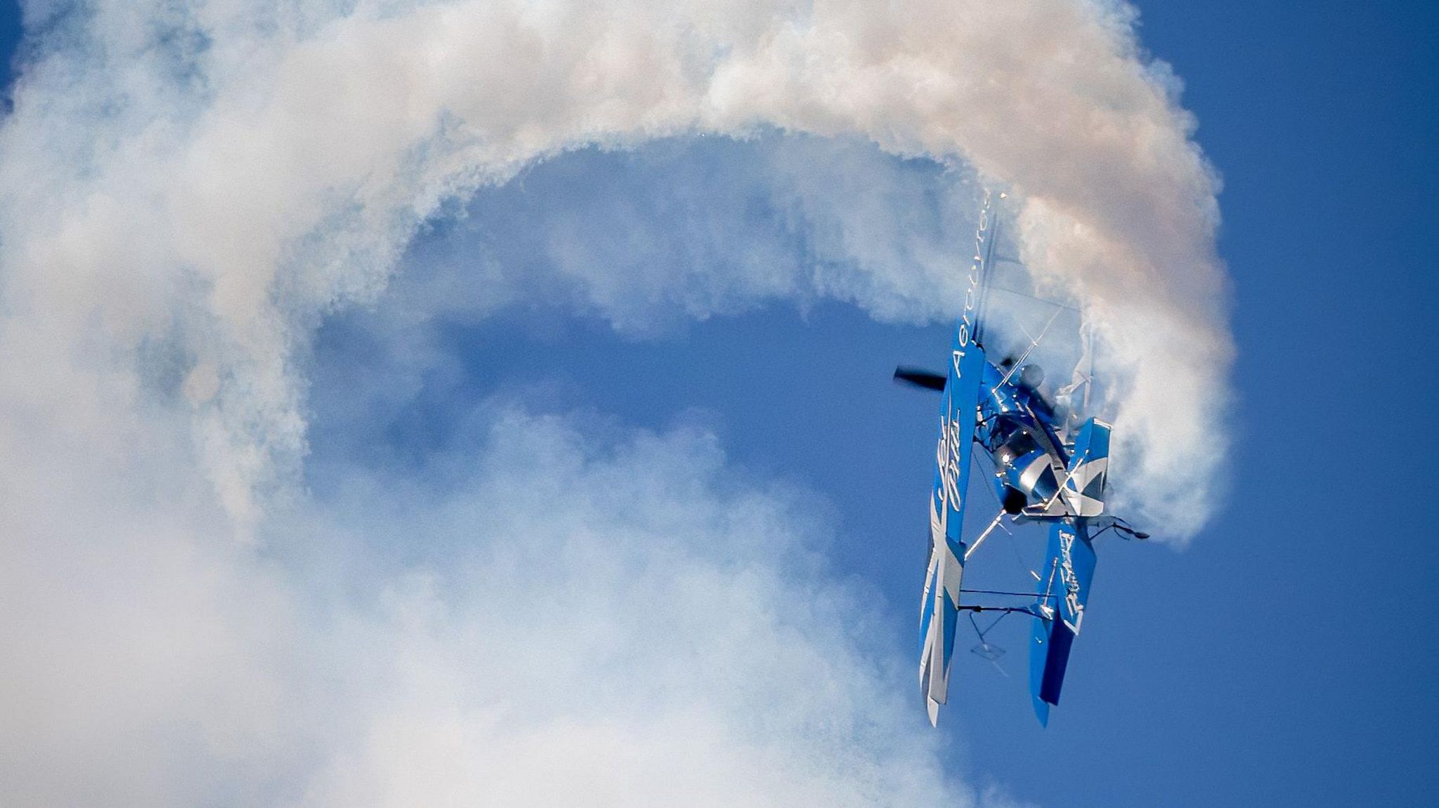 A blue and white plane performs a barrel roll and leaves a plume of white smoke behind it