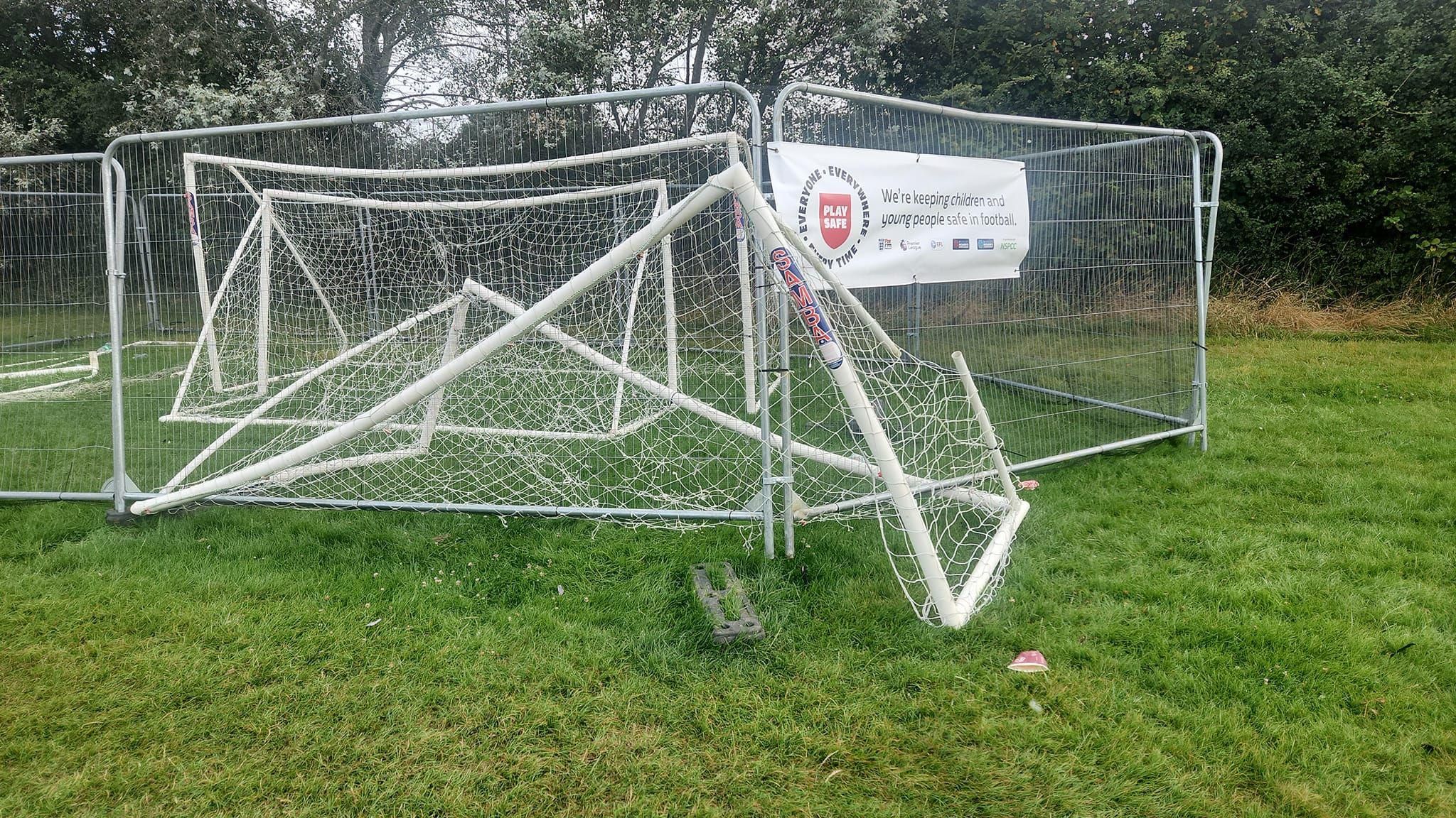 A plastic goal half under tall fencing, bent over in a field with trees at the edge