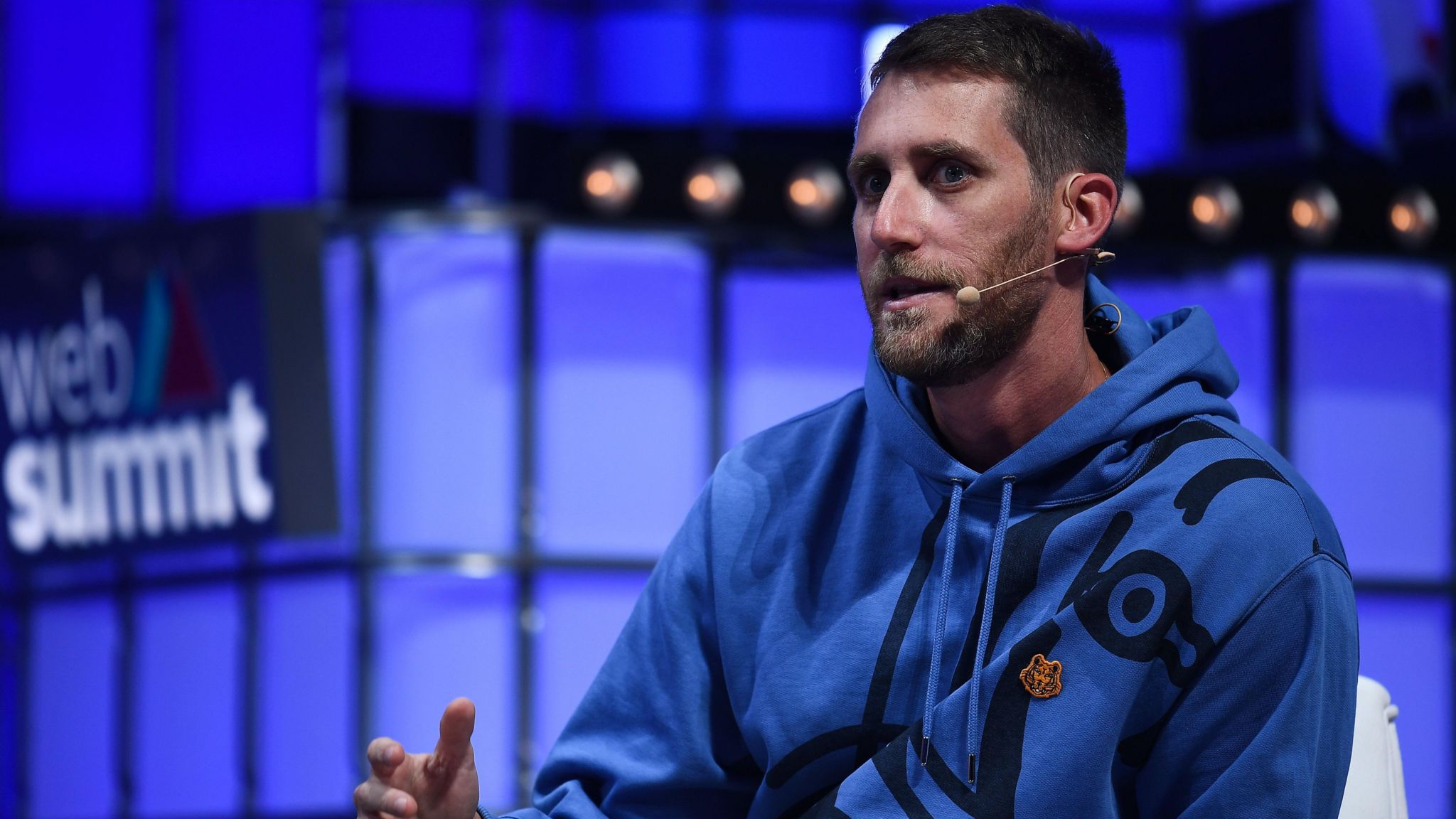 A man speaking on-stage at an event. He's wearing a blue hoody and has a smart beard and hairstyle