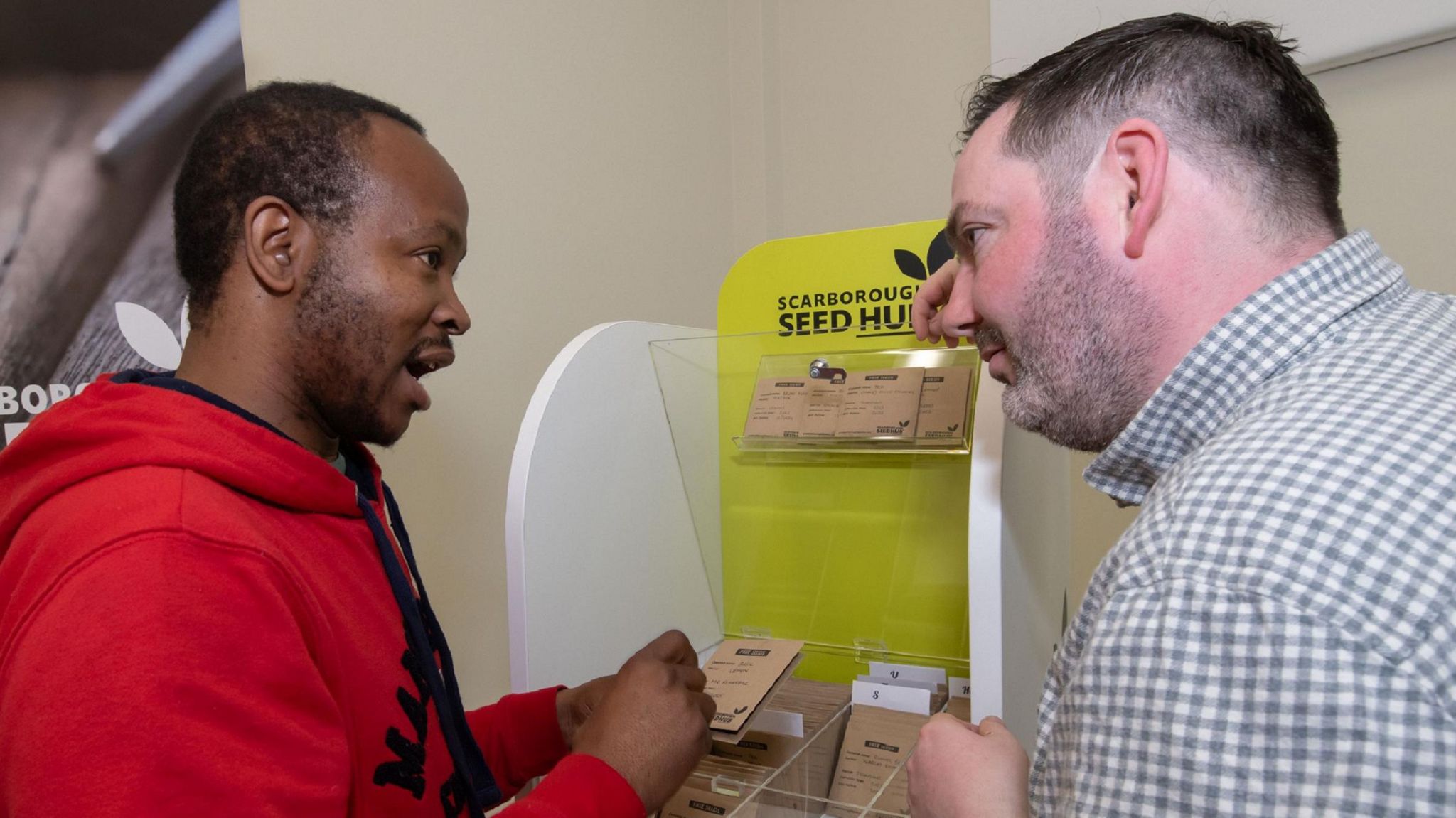 The co-chairman of Grow Scarborough, Darren Mancrief, is pictured in front of a desk containing seeds in brown packets. He is showing these seeds to a visitor Athenkosi Nyengane