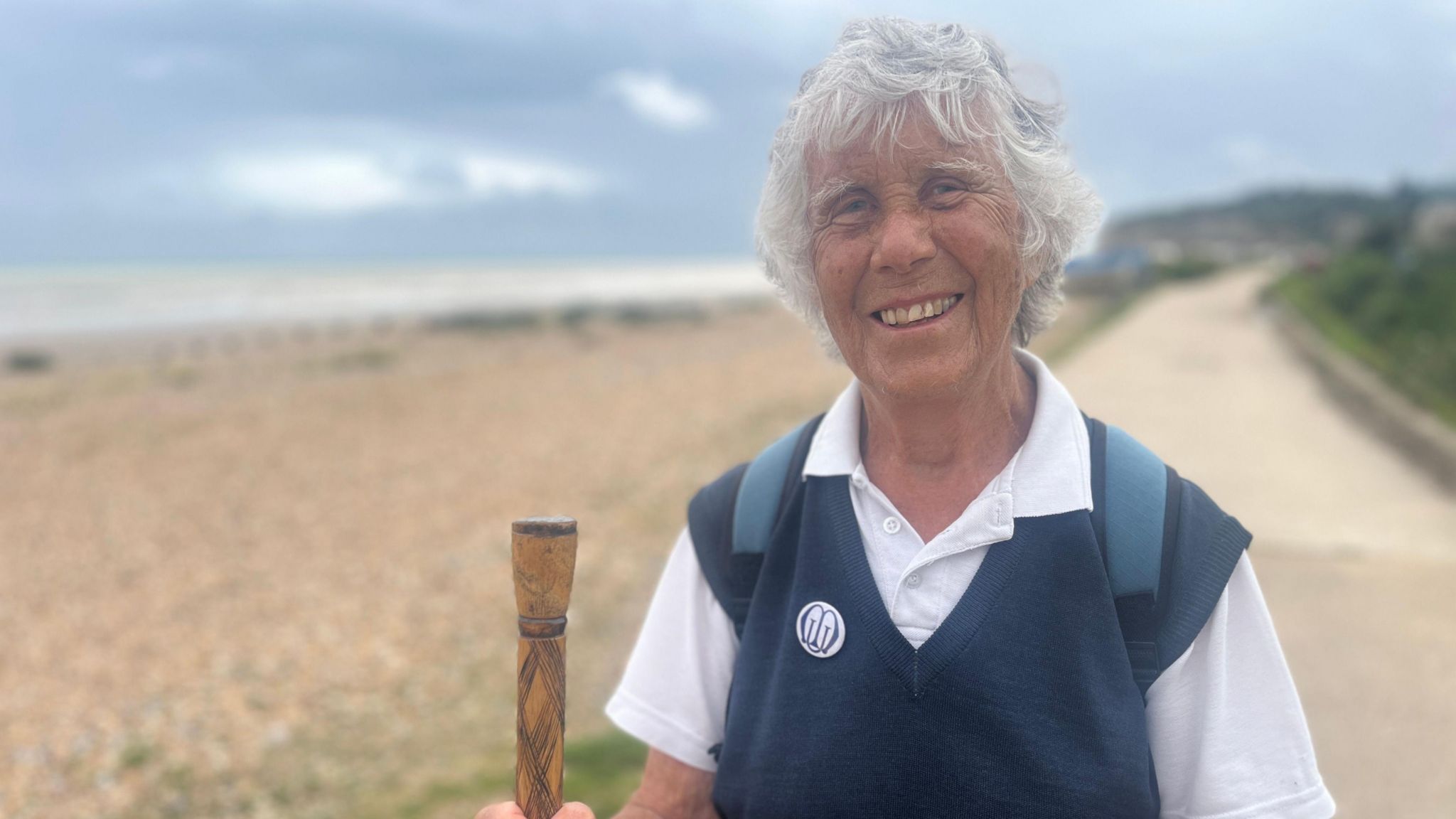 Pam Brooks, a grey haired woman, stands with walking stick, on a coastal path, smiling at the camera