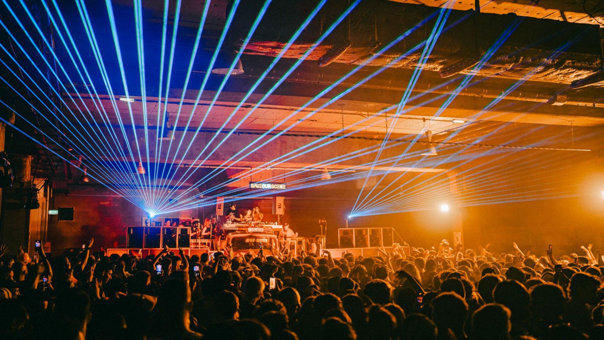 An underground rave with a lot of people in the crowd, lasers and a Save Our Scene lighbox in the backgroud