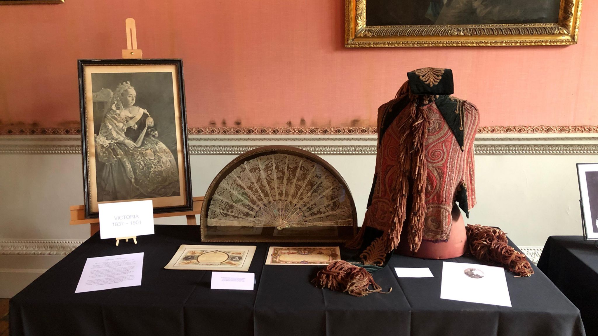 A portrait of Queen Victoria, a hand fan and a maid in waiting uniform on top of a table