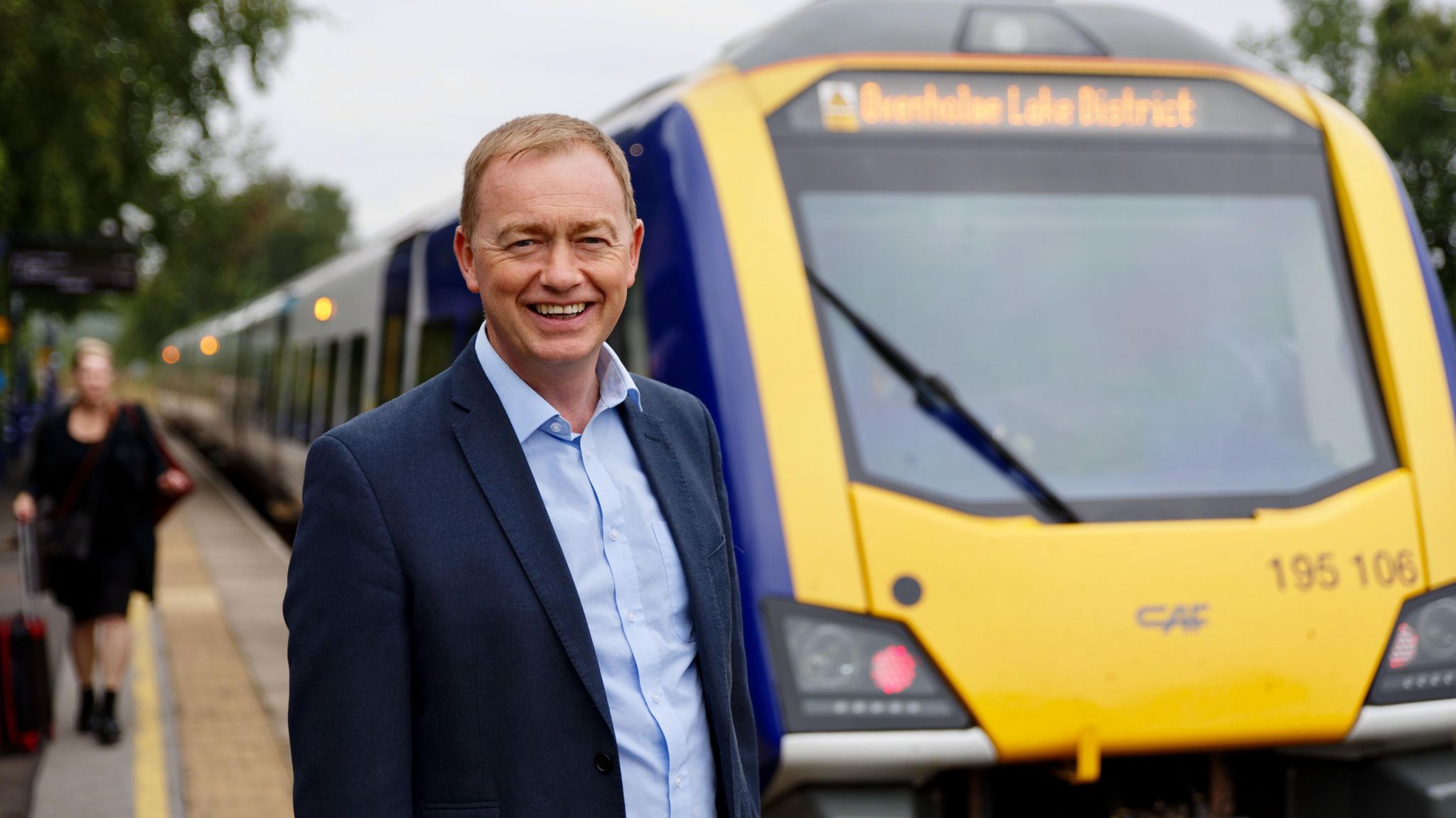 The MP Tim Farron, wearing a blue suit, stands in front of a train which has the words Lake District on the front.