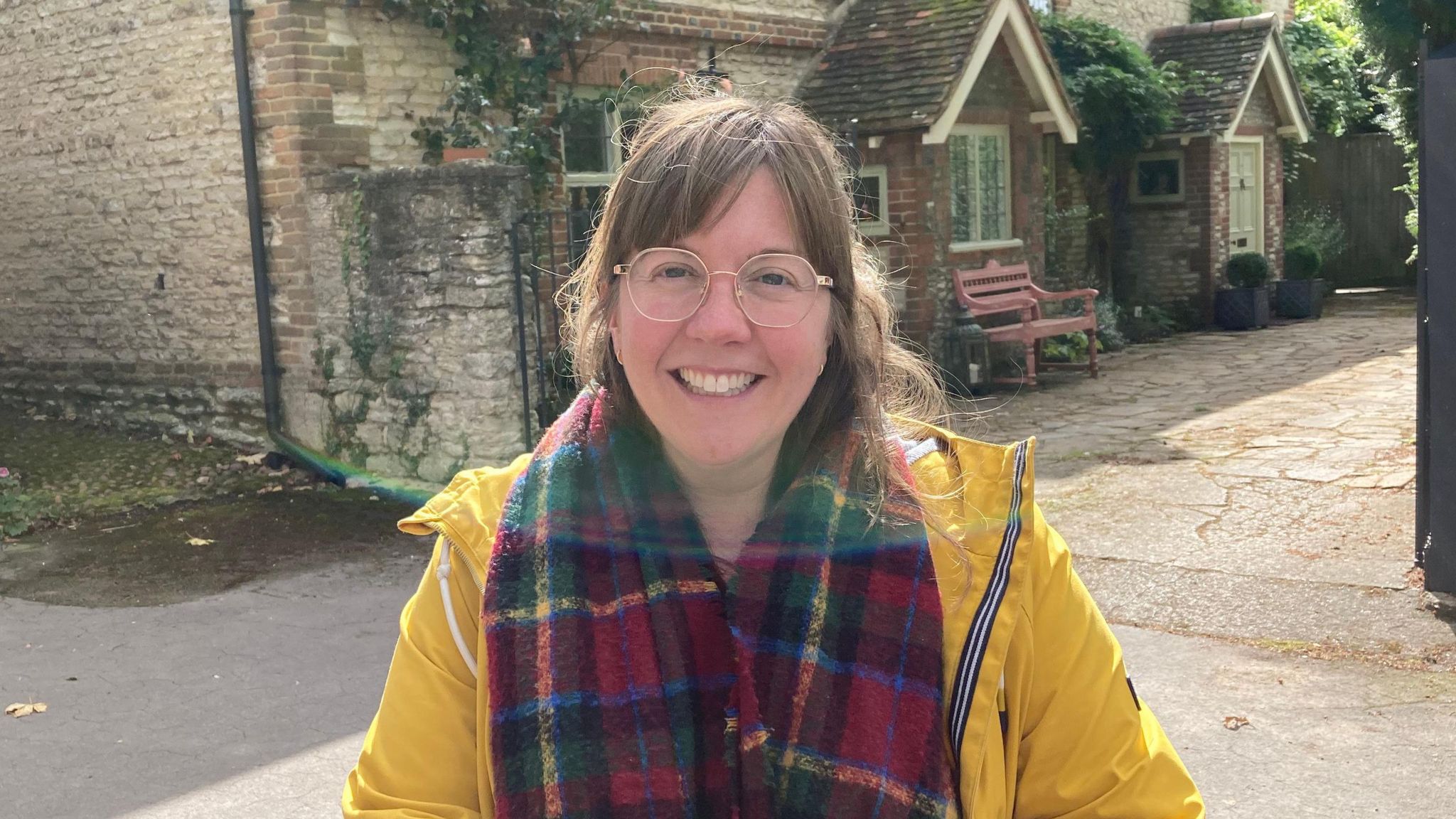 Sarah Mason from the sternum up. She has long-ish brown hair, glasses and is wearing a bright yellow coat. An old building is in the background