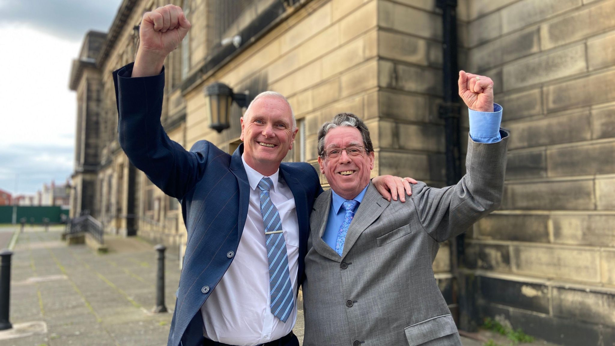 Keith Marsh, wearing a navy blue suit jacket, white shirt and blue striped tie and Robin Clarke, wearing a grey suit and light blue shirt and tie stand together, smiling and raise their fists in the air outside the town hall 