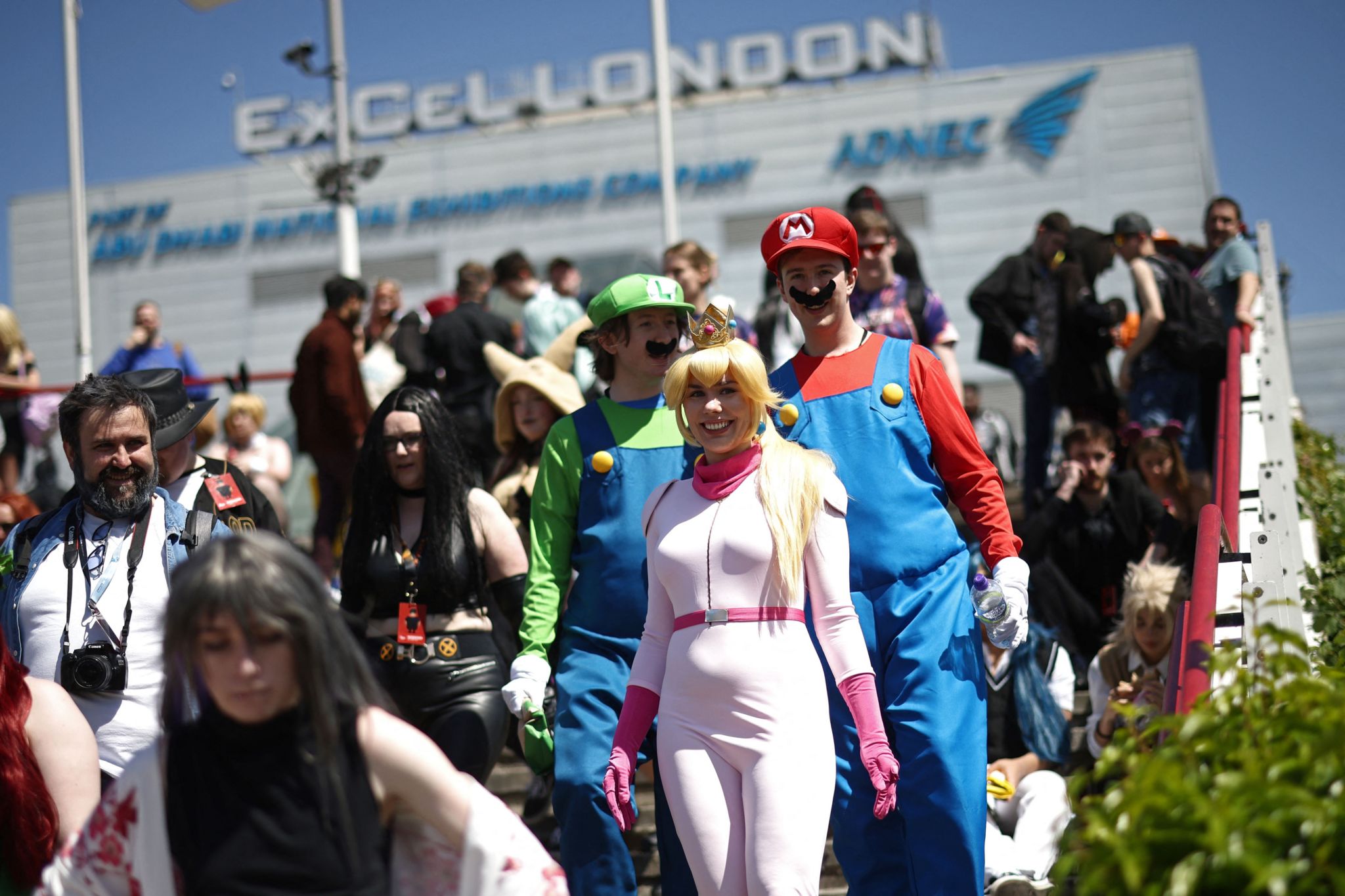 Outside of ExCel in London where Comic Con is held. The picture features people attending the convention, dressed up as various characters. The main people in the photo are seen dressed as characters from Super Mario Bros. A woman is dressed as Princess Peach, wearing a blonde wig, a gold crown and a light pink jumpsuit with dark pink gloves. Two men behind are dressed as Mario (right) and Luigi (left). Both men are wearing blue overalls and dark brown moustaches. Luigi is wearing a breen shirt underneath his overalls and a green hat with the letter "L" on it. Mario is wearing a red shirt under his overalls and a red hat with the letter "M" on it. Other people in various costumes surround them.