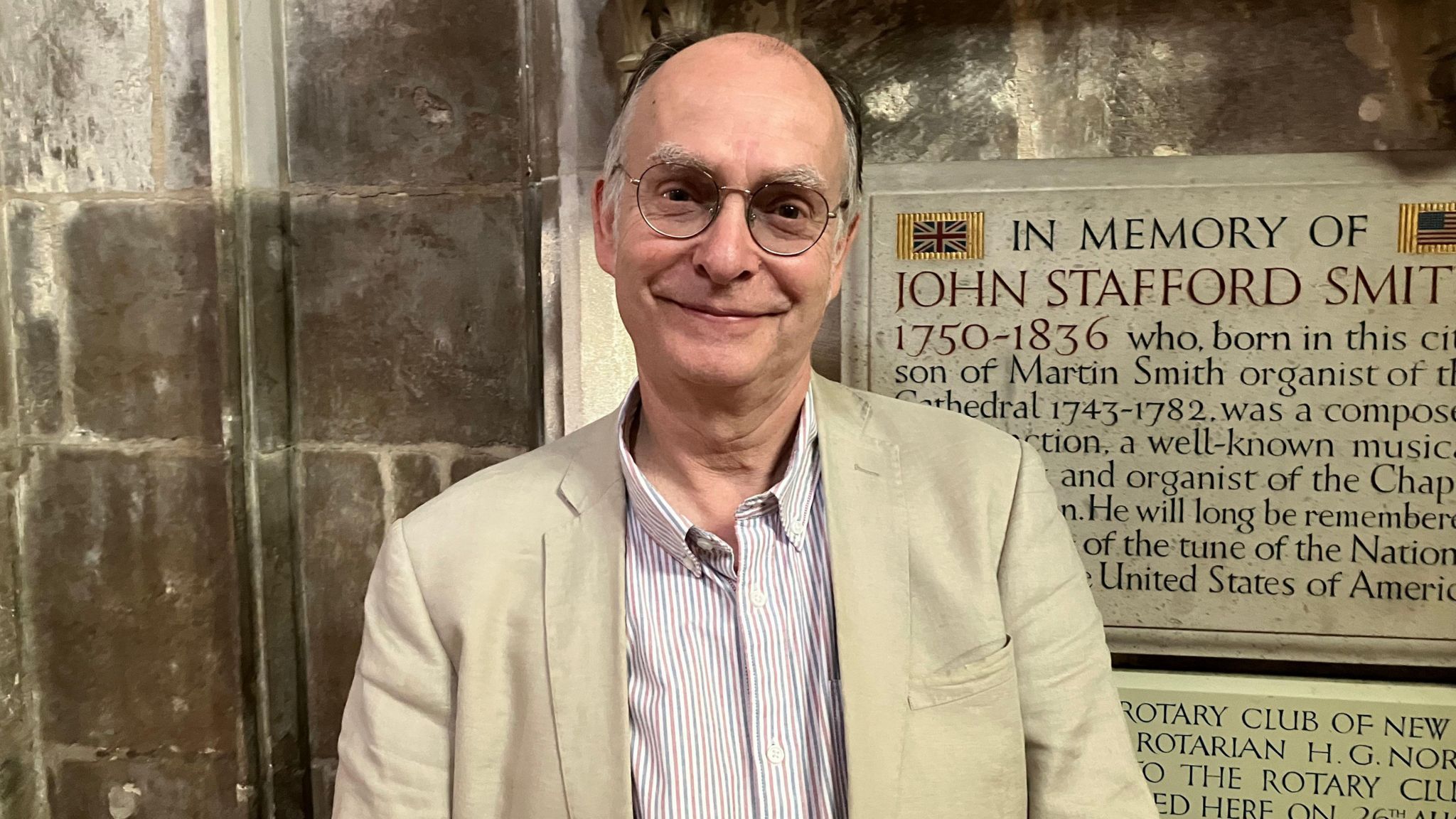 Adrian Partington wearing a striped shirt and glasses. He is smiling and standing in front of a plaque dedicated to John Stafford Smith