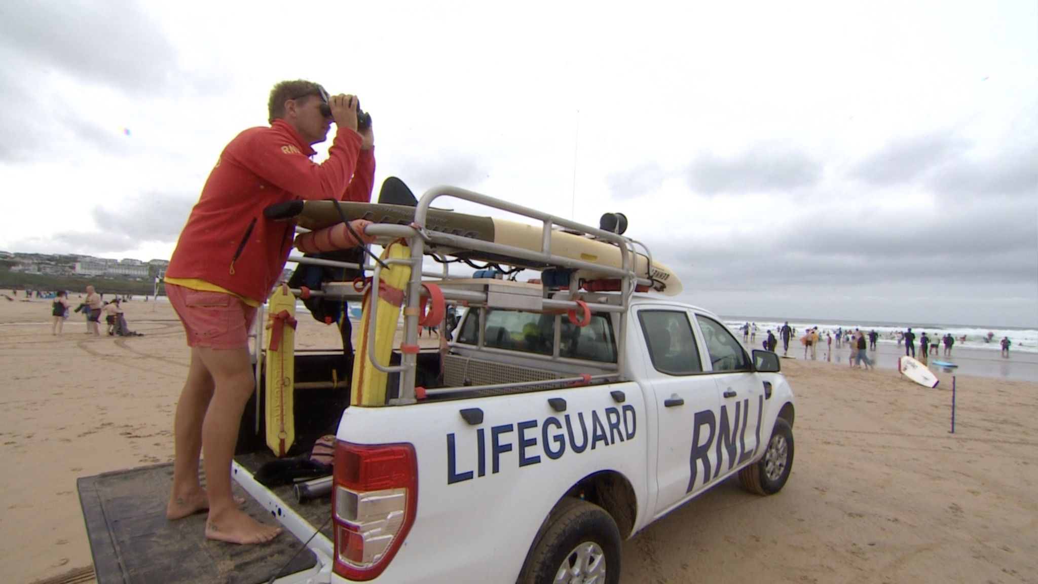 A busy beach with choppy waves. An RNLI Lifeguard truck with a lifeguard standing on the back looking out to sea with binoculars. 