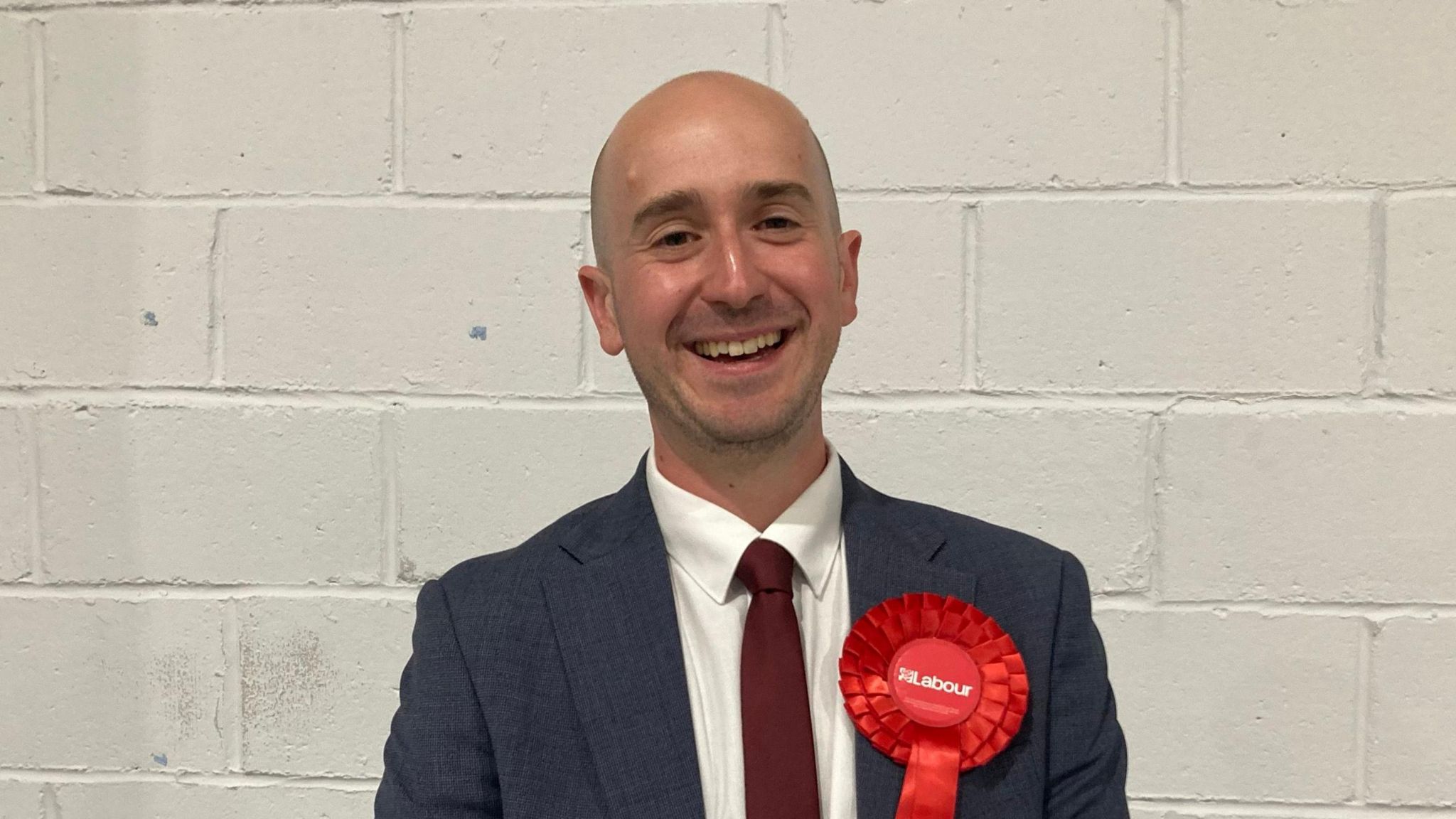 A smiling Ben Goldsborough looks directly at the camera dressed in a dark suit and tie, with a red Labour rosette attached to his lapel