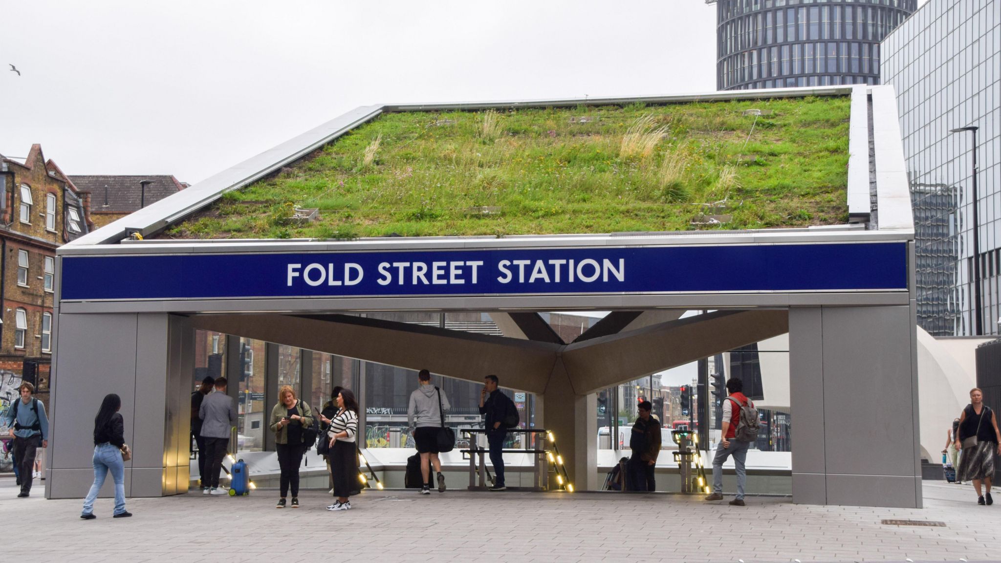 Passengers going in and out of the entrance to an underground station, with a large blue sign above the archway. Written on the sign in white capital letters are the words "Fold Street Station".