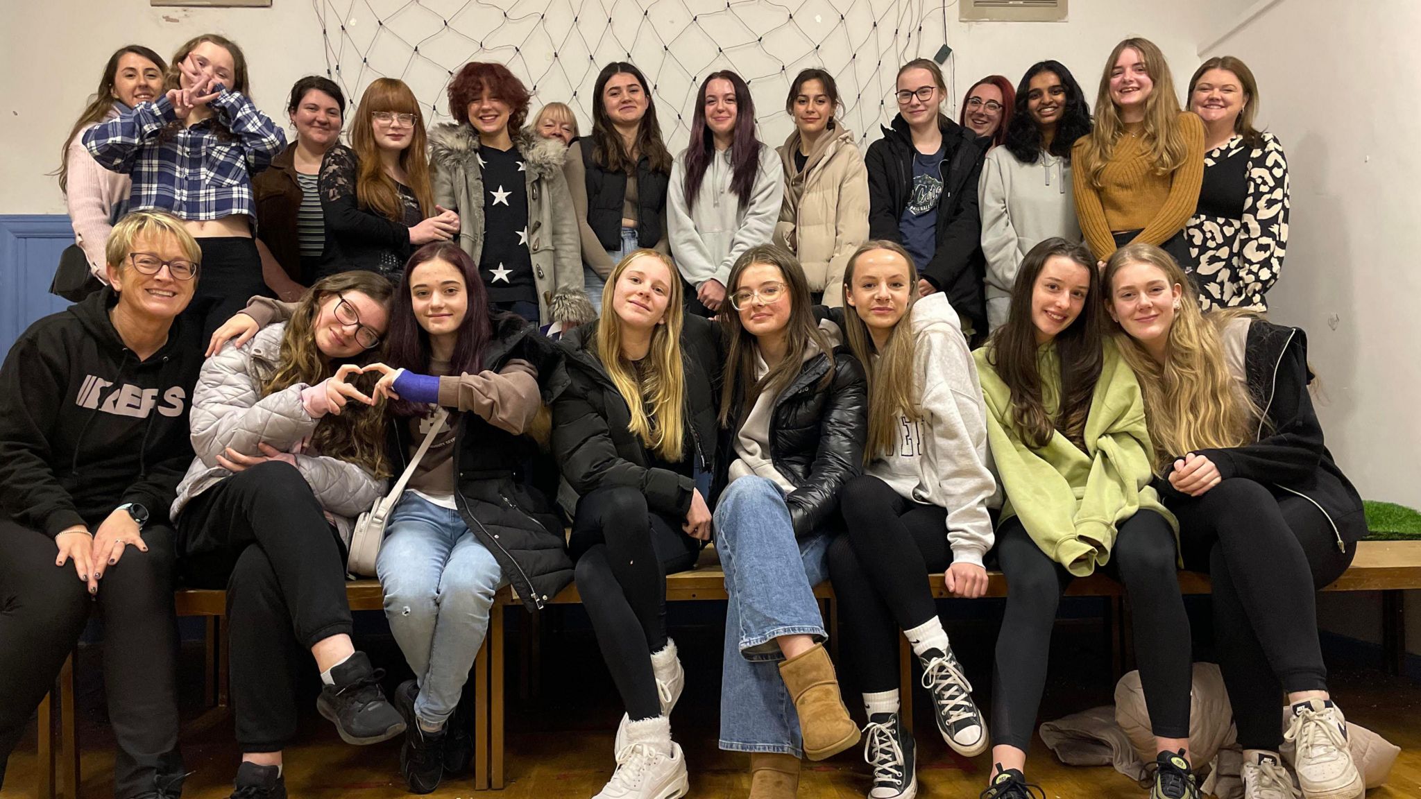 Members of the United Youth Alliance - 22 young girls and United Youth Alliance workers - dressed in hoodies and coats smile at the camera in two rows. One holds up peace signs. 