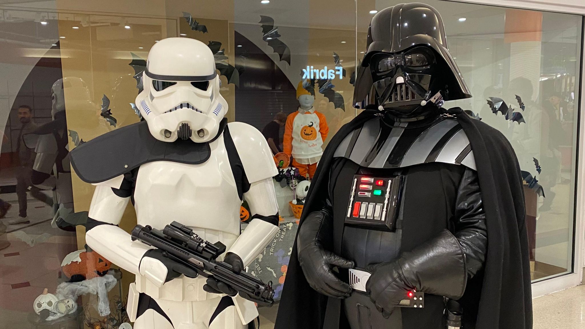 Two people, one dressed as Darth Vader and one as a Stormtrooper, pose for a photo in front of a shop window. 