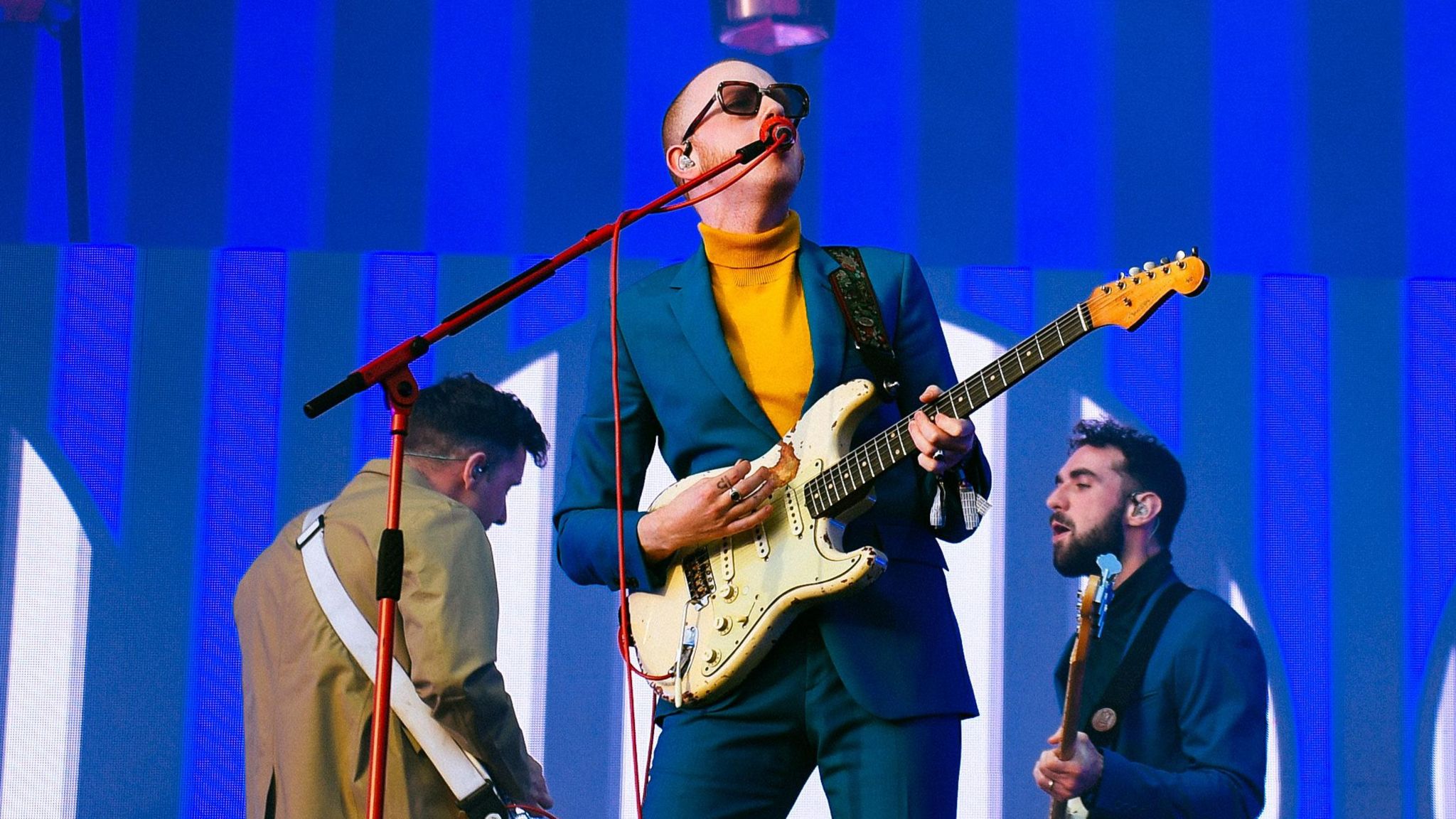 Two Door Cinema Club performing. Lead singer Alex Trimble is playing guitar and singing into a microphone.