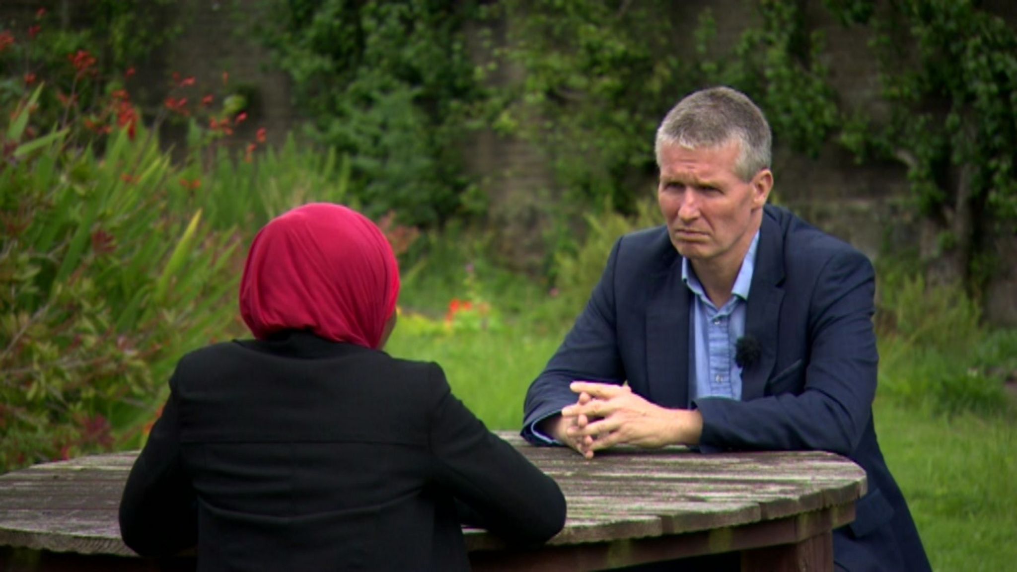 A green garden with a wooden picnic table and Faiza with her back to the camera, sitting opposite the reporter. She is wearing a bright pink hijab and black blazer. The correspondent is sitting facing Faiza and the camera. He is looking pensive with his hands crossed. He is wearing a blue shirt and navy suit. 