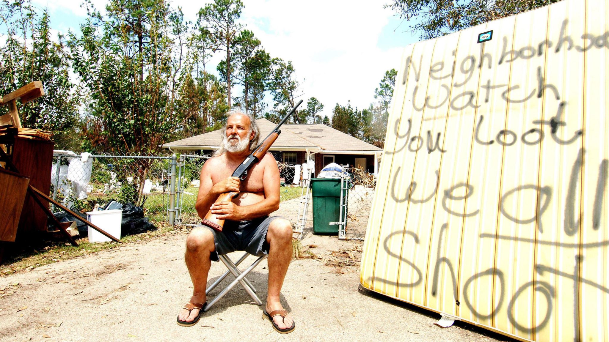 A man sits in shorts and flip flops, holding a gun. He is next to a sign which reads "neighbourhood watch you loot we all shoot".