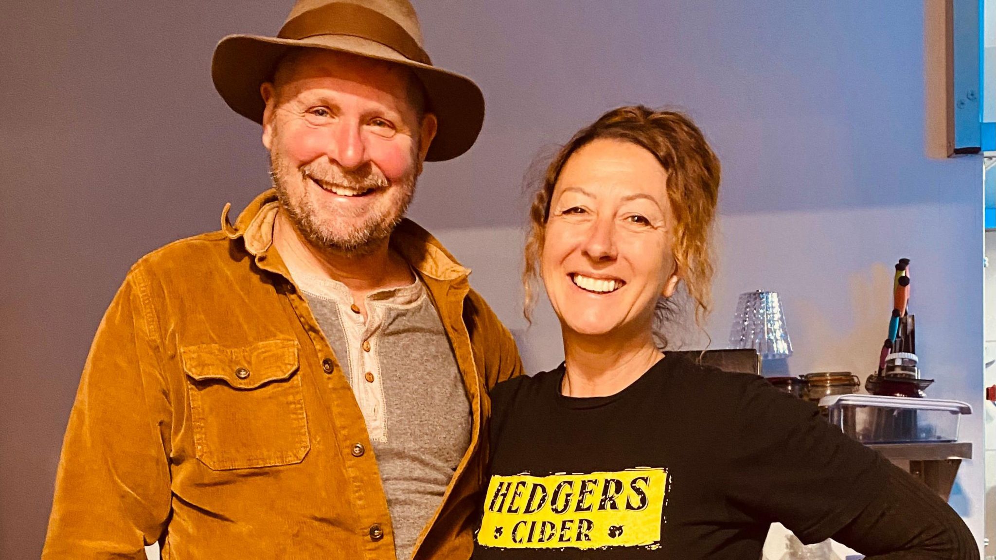 Tom and Jo standing side by side smiling at the camera. Tom is wearing a grey t-shirt, light brown jacket and brimmed hat. Jo has her hair tied up and is wearing a black t-shirt with a yellow Hedgers Cider logo across the chest