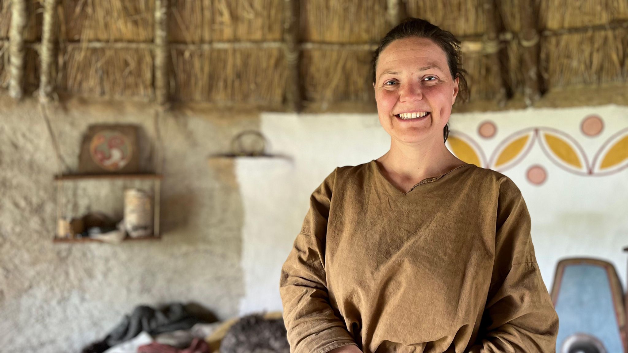 A woman smiling to camera, who is wearing a brown top and stood inside an iron age roundhouse, with paintings on the wall behind and the bottom of a thatched roof visable.