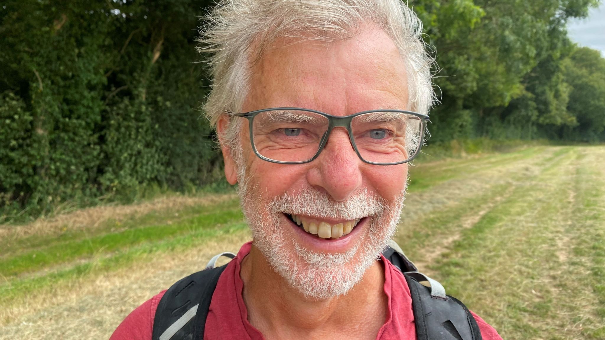 An older man with white hair and beard and glasses smiling at the camera, with a field and trees in the background