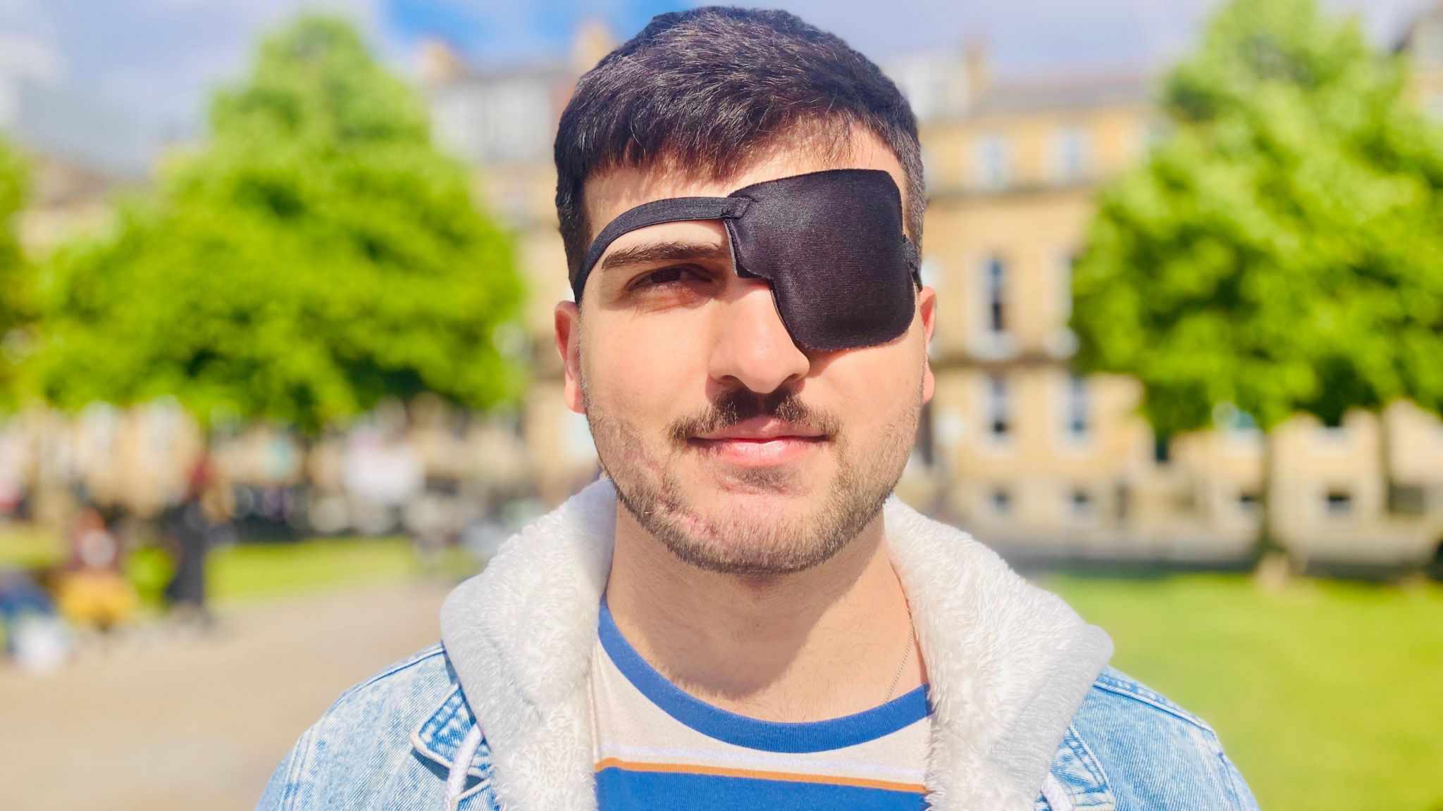 Jabar Barzanji - a man with dark hair has a patch over his left eye. He wears a denim jacket and stands in front of a small park