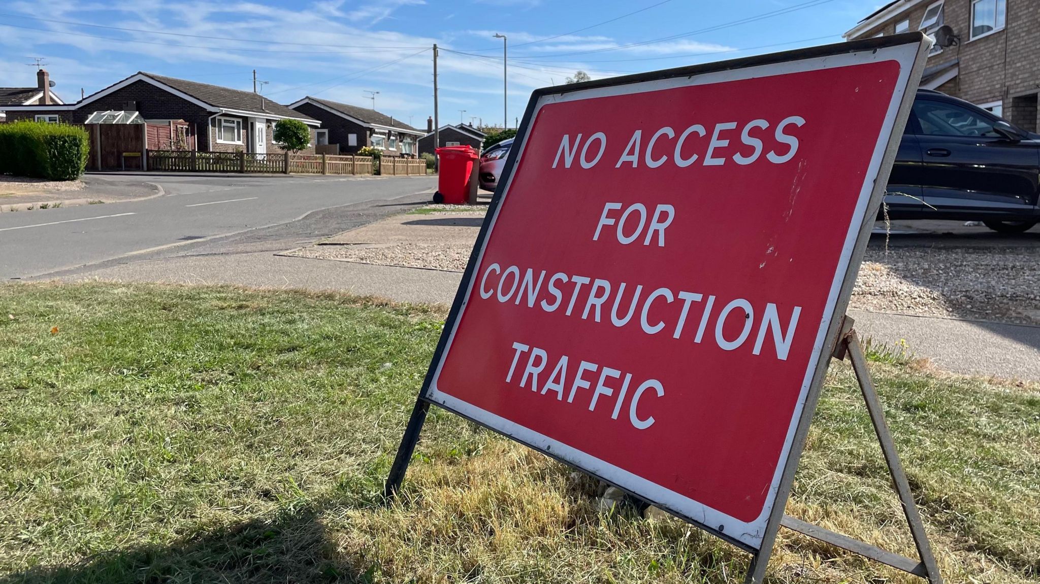 A temporary rectangular roadsign that reads "no access for construction traffic" in white text on a red background. It is standing on a grass verge by the side of the road. Two bungalows can be seen in the background.