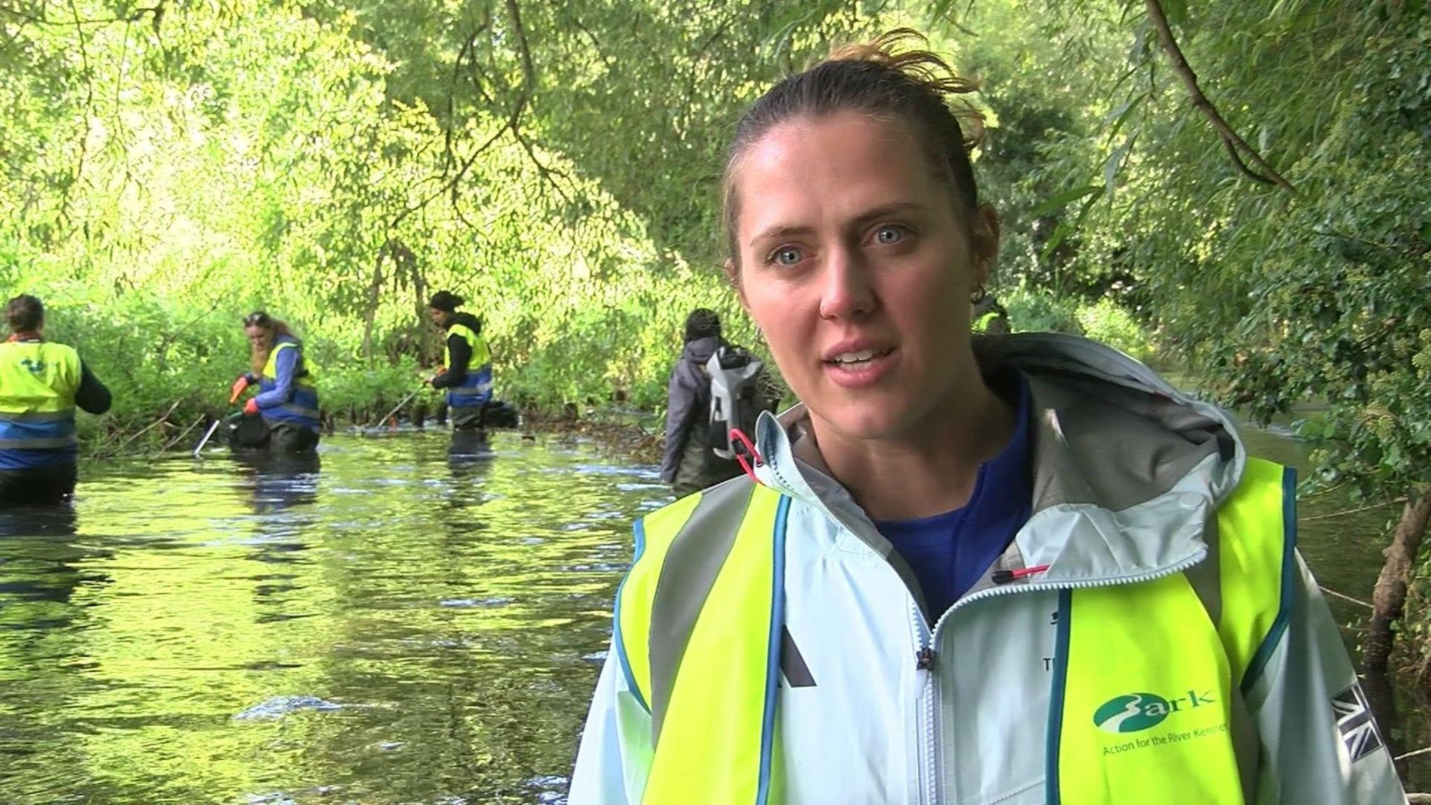 A woman with blonde hair tied up in a bun, wearing a blue waterproof jacket and a high-visibility vest saying 'Action for the River Kennet' on it. She's standing in front of a river with lots of trees around it, with three people wearing waders and high-visibility jackets picking up litter from the water.