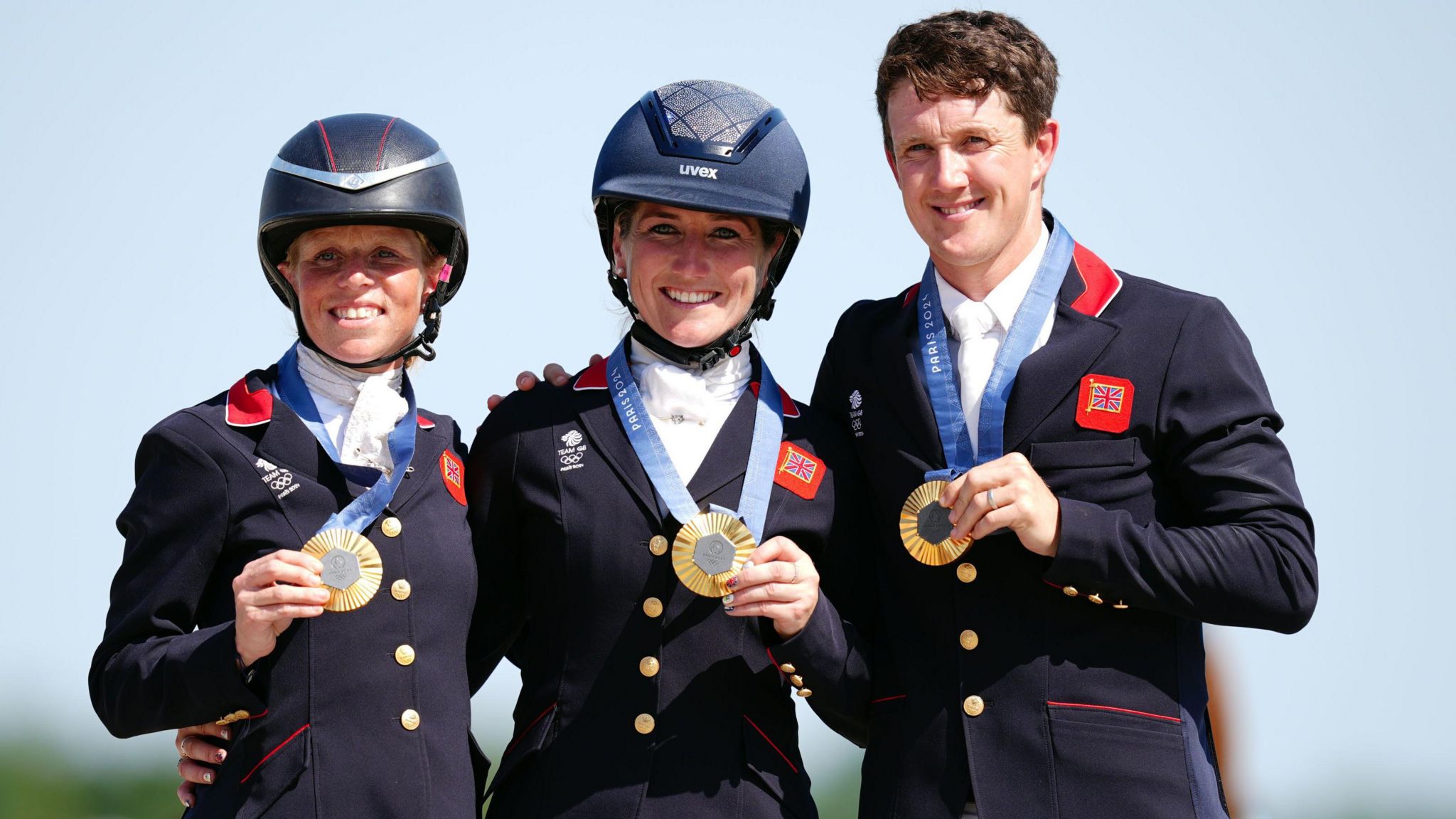 Rosalind Canter, Laura Collett and Tom McEwen. Rosalind and Laura are wearing horse riding helmets, whilst Tom is not. The trio are all dressed in equestrian gear and wearing medals around their necks. They have their arms around each other and are smiling 