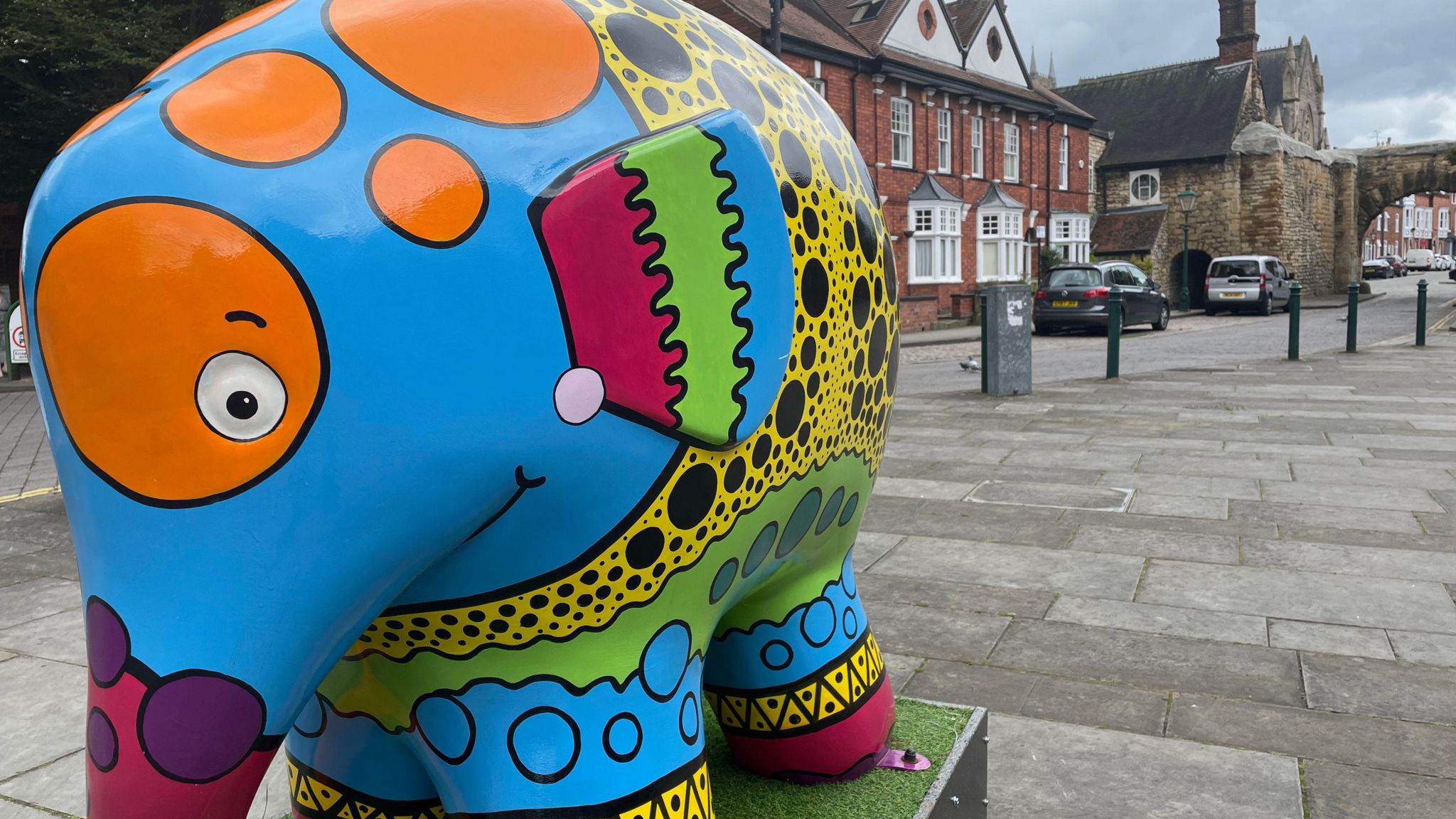 A statue of Elmer the elephant standing on a plinth on the street and is a vibrant mix of bright colours with an orange patch around his eye and yellow and black spots on his back