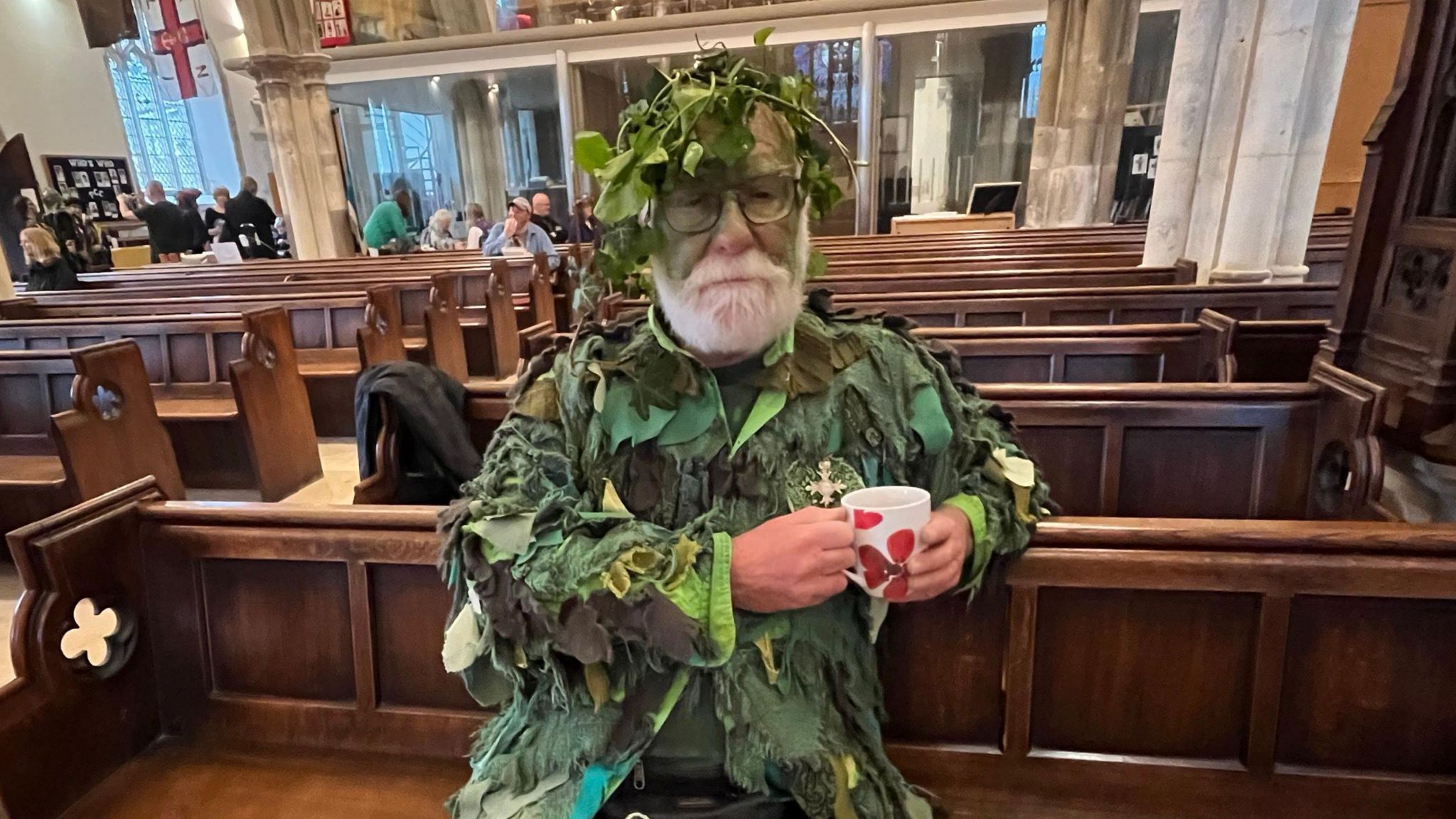 An older man with a white heard is painted in emerald green and wears a multi-shade green costume drinks a cup of tea in a church pew