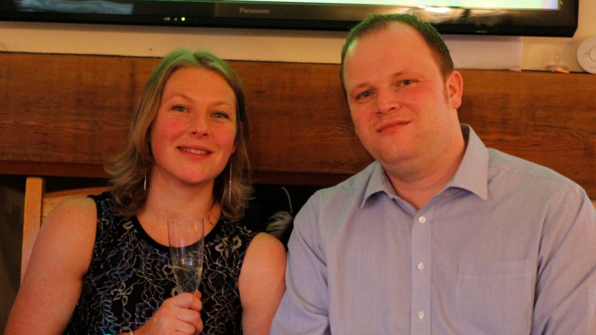 Alison and Rob Clark pose for a photo together. She is smiling, wearing a black top with gold pattern and holding a champagne flute. He is wearing a light blue shirt.