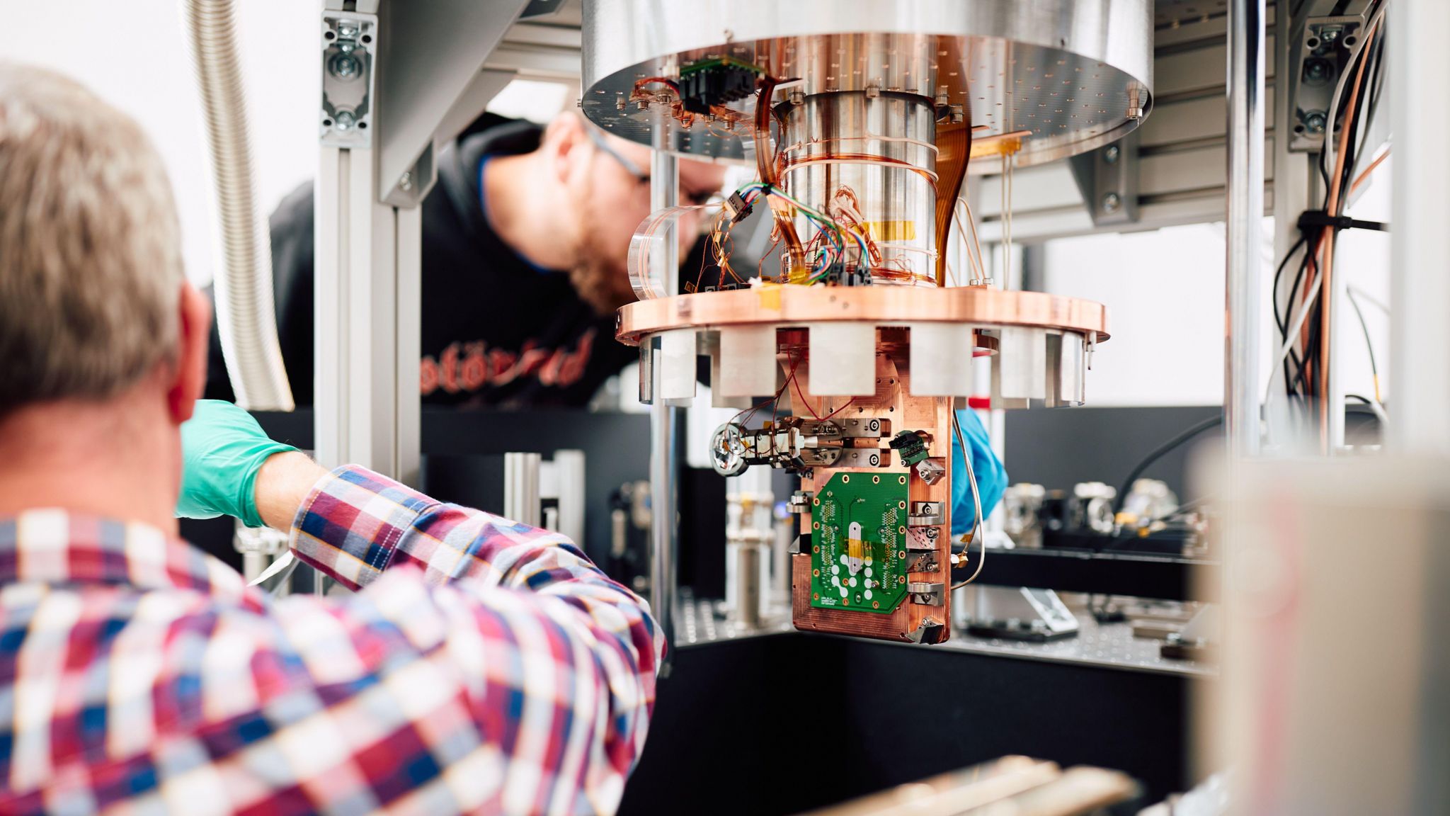 A green computer chip attached to a large copper coloured fixture with two men, both wearing gloves, working on it